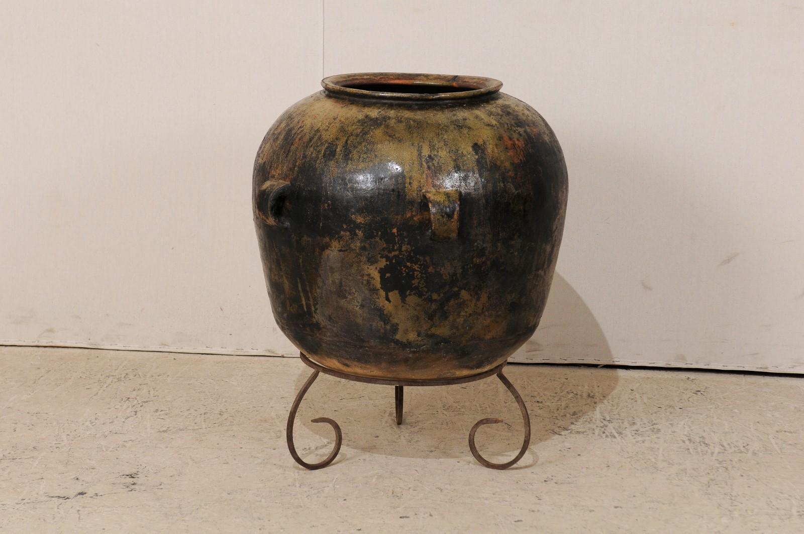 A Guatemalan ceramic jar on stand. This Guatemalan jar, from the early 20th century, features a wide body, four handles, and has a nice patina with rich warm hues throughout. The jar is raised on a custom iron tripod base with scrolled feet.