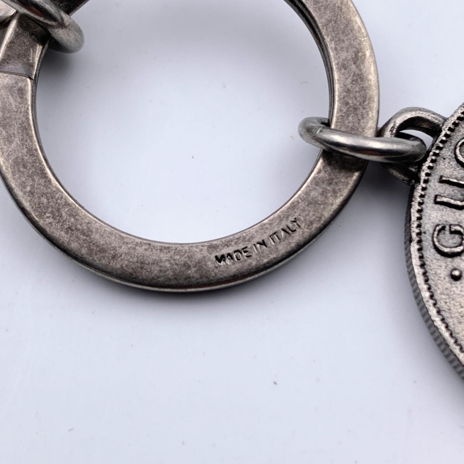 Gucci keyring in aged silver metal. Round coin charm with embossed tiger head with 'L'Aveugle Par Amour -1921' on front. Gucci logo with number 25, 2 bees, and the 'Loved' writing on the back. Gucci engraved on clasp and ring. Total length: 4.5