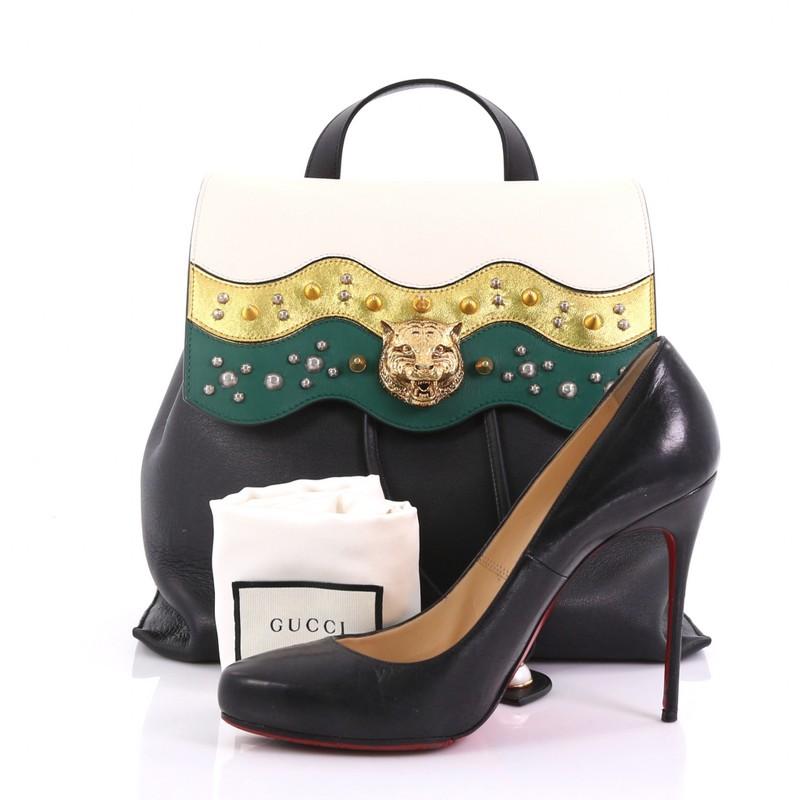 This Gucci Animalier Malin Backpack Studded Leather Medium, crafted in black leather, features studded wave-pattern flap in white, gold, and green, a statement feline head at the center, gold-tone chain straps, and gold-tone hardware. Its hidden