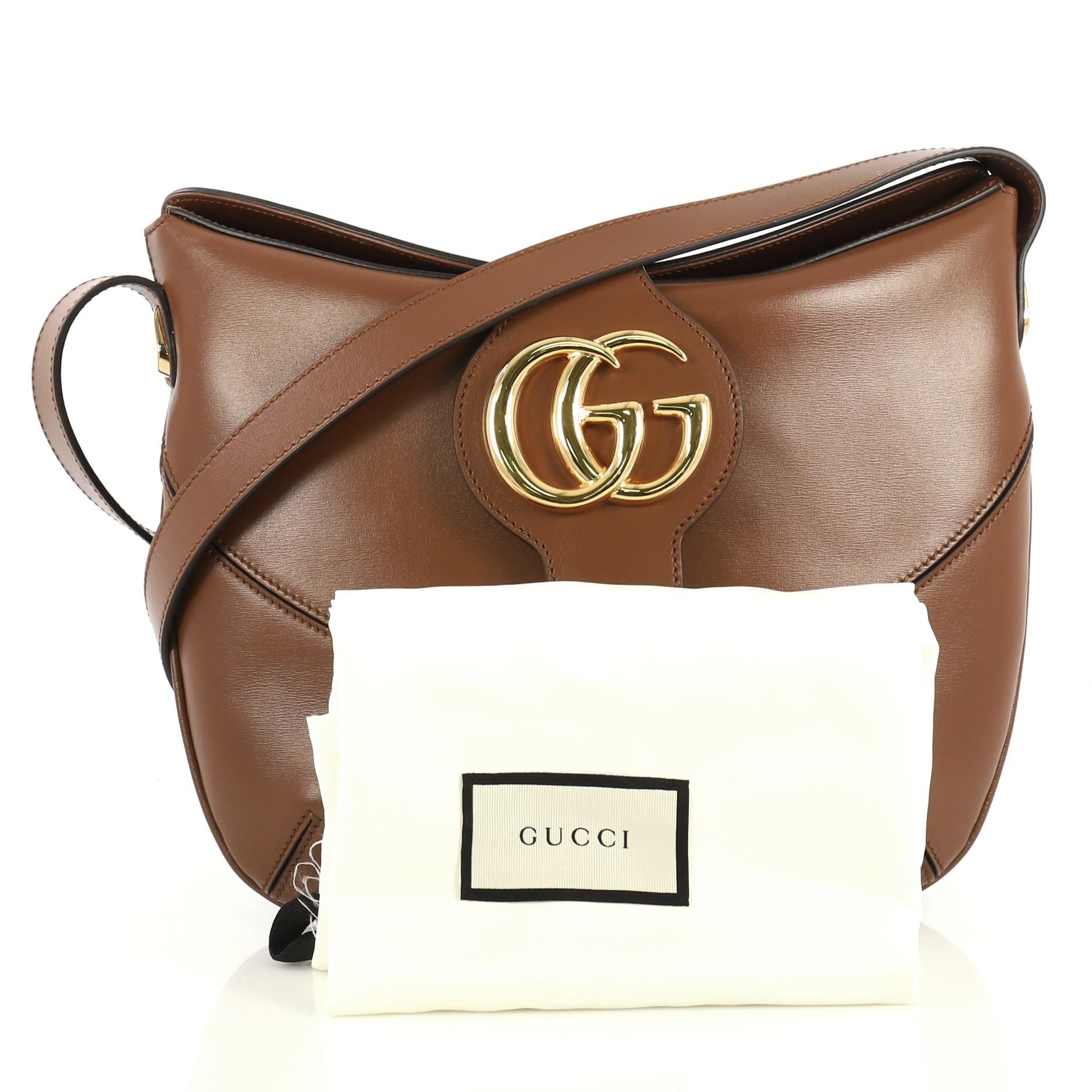 This Gucci Arli Crossbody Bag Leather Medium, crafted from brown leather, features an adjustable leather strap, flap top with GG logo, and gold-tone hardware. Its magnetic snap closure opens to a brown microfiber interior with zip and slip pockets.