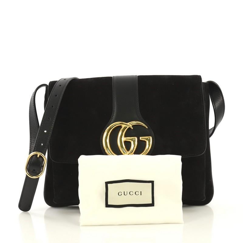 This Gucci Arli Shoulder Bag Suede with Leather Medium, crafted from black suede with leather, features an adjustable leather strap, flap top with GG logo, and aged gold-tone hardware. Its magnetic snap closure opens to a pink fabric interior with