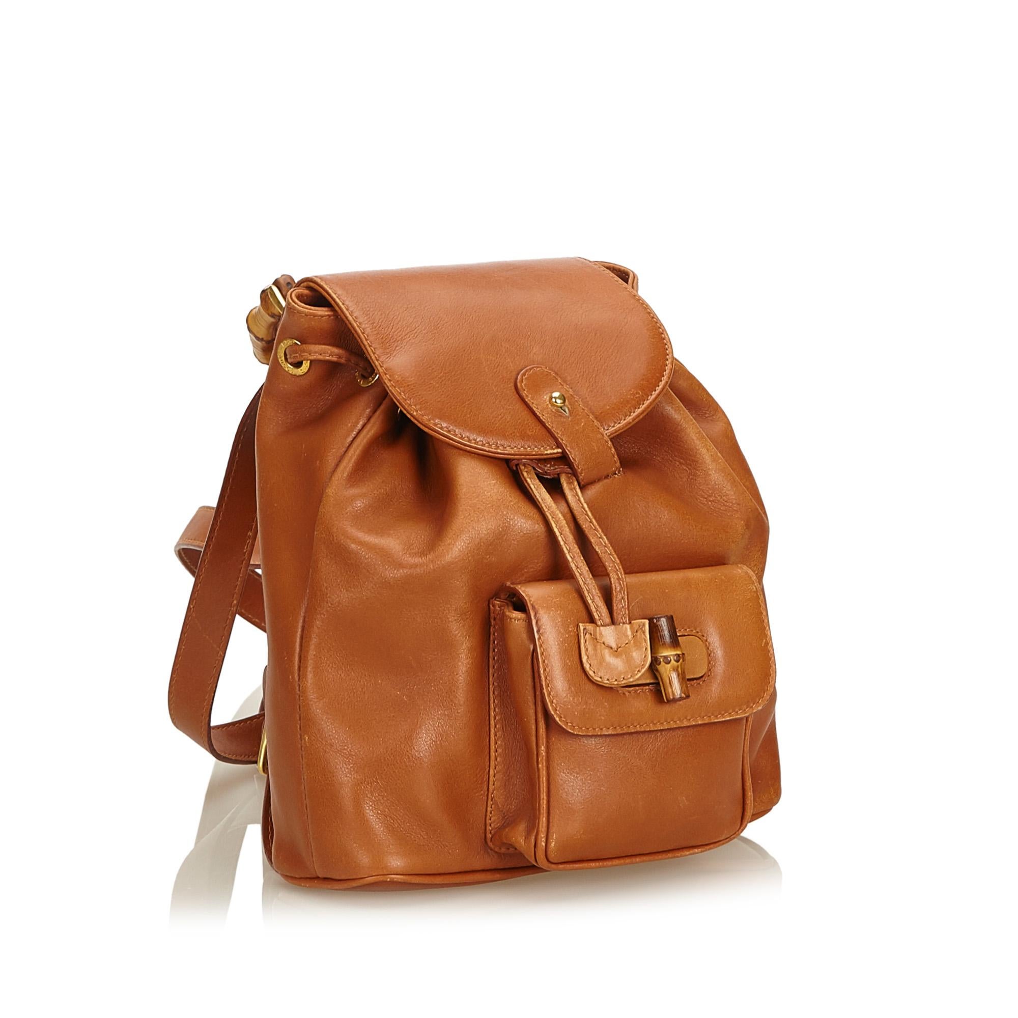 This vintage Gucci mini backpack features a tan leather body, flat back straps, bamboo top handle, top flap, drawstring closure, exterior flap pocket with bamboo twist lock closure, and interior zip pocket.