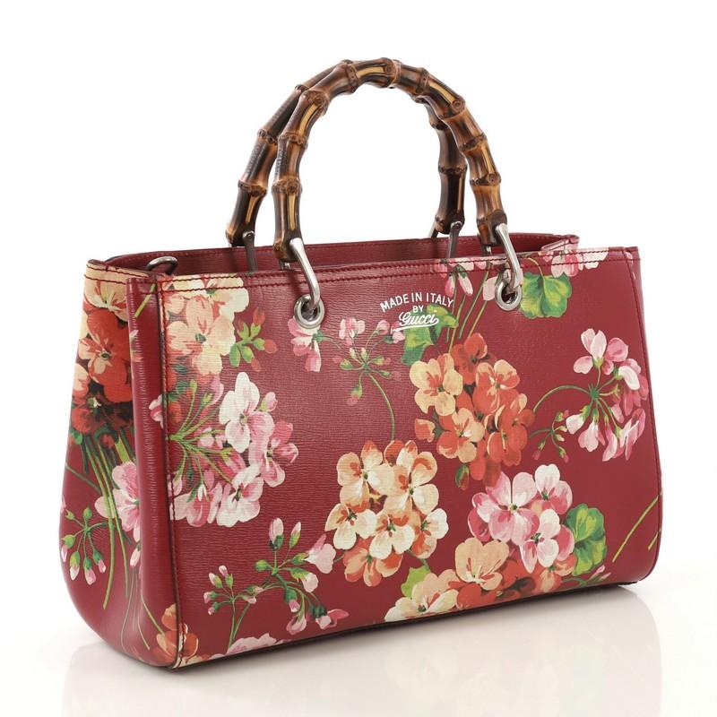 This Gucci Bamboo Shopper Tote Blooms Print Leather Medium, crafted from red blooms print leather, features sturdy bamboo handles, protective base studs, stamped logo at the front, and aged silver-tone hardware. Its hidden magnetic snap closure