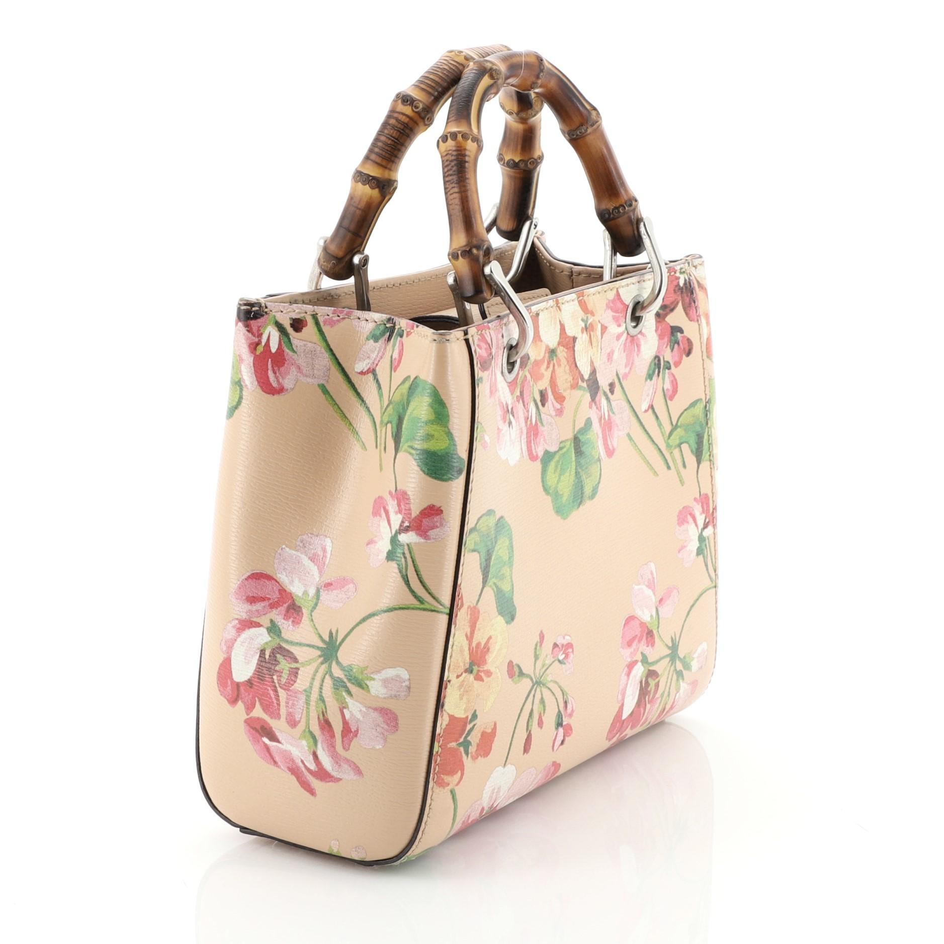 This Gucci Bamboo Shopper Tote Blooms Print Leather Mini, crafted from pink blooms print leather, features dual bamboo handles, stamped logo at the front, and aged silver-tone hardware. Its hidden magnetic snap closure opens to a neutral canvas