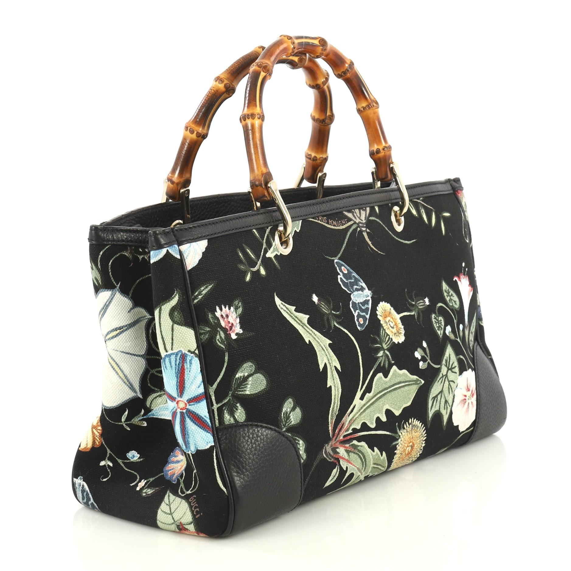This Gucci Bamboo Shopper Tote Flora Canvas Medium, crafted from black printed canvas, features dual bamboo handles, floral motif, leather trim, and gold-tone hardware. Its magnetic tab closure opens to a neutral fabric interior with middle zip