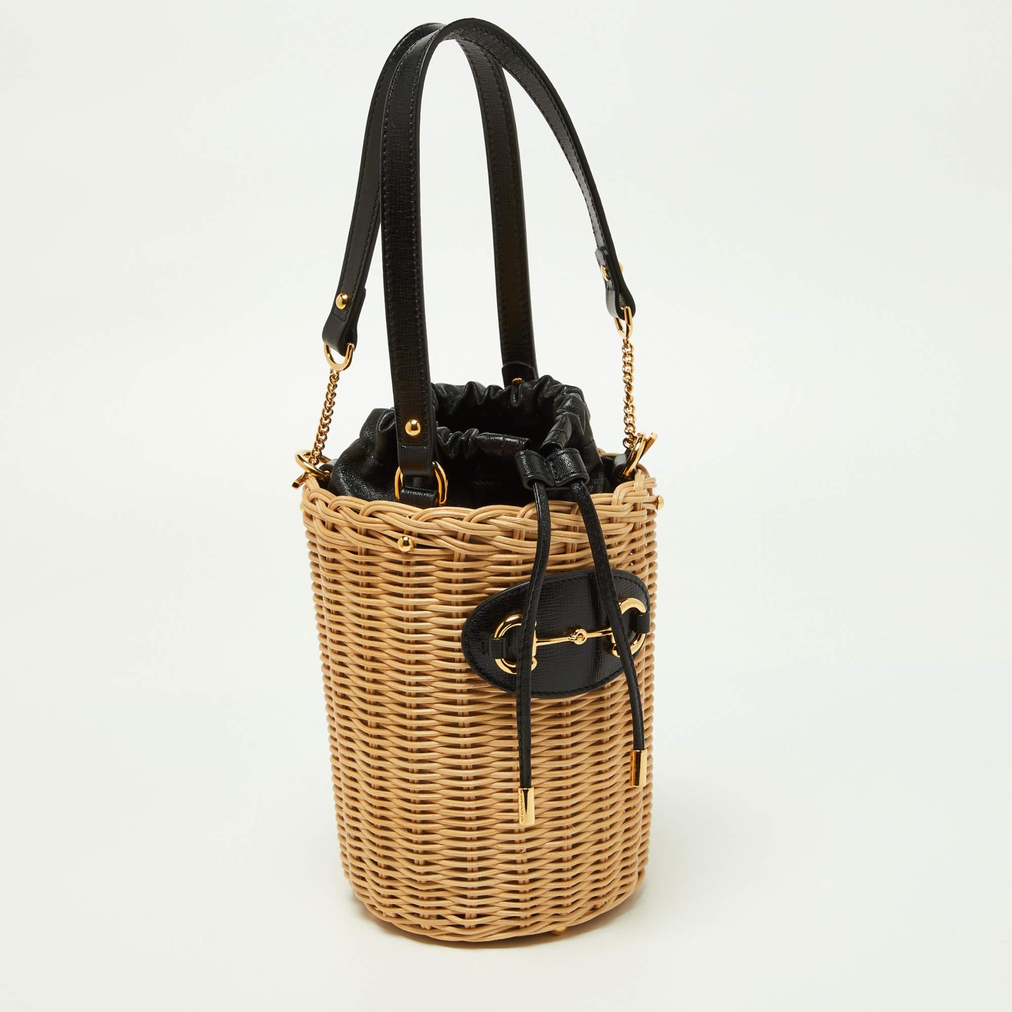 The Gucci Horsebit 1955 bucket bag is a chic and versatile accessory. Its body features beige wicker with black leather trim, adorned with the iconic Horsebit detail. The bag has a spacious interior, drawstring closure, and an adjustable leather