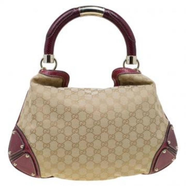 Die Tasche ist aus beigem Canvas mit GG Monogrammen gefertigt und hat einen roten Kontrastbesatz. Ein steifer, gerollter Griff erleichtert das Tragen, und die Lederecken ergänzen das Design.  Modisch übertriebene Lederquasten und die