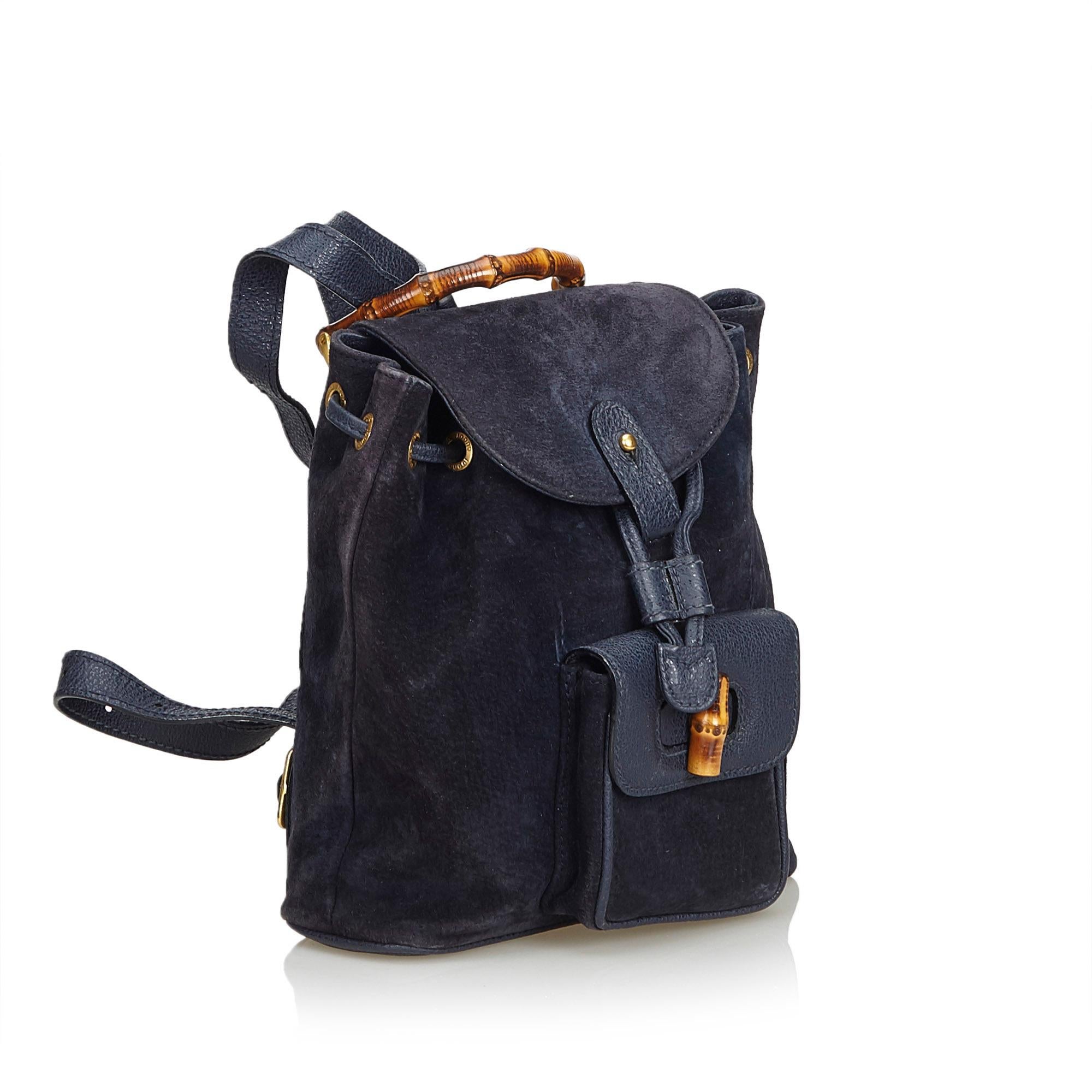 This backpack features a suede body, flat leather back straps, bamboo top handle, top flap with button closure, drawstring closure, exterior flap pocket with bamboo twist lock closure, and an interior zip pocket. It carries as B+ condition