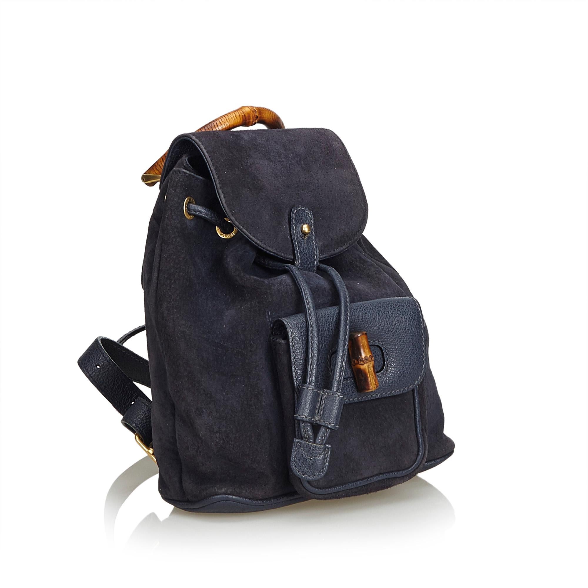 This backpack features a suede body, flat leather back straps, bamboo top handle, top flap with button closure, drawstring closure, exterior flap pocket with bamboo twist lock closure, and an interior zip pocket. It carries as B+ condition