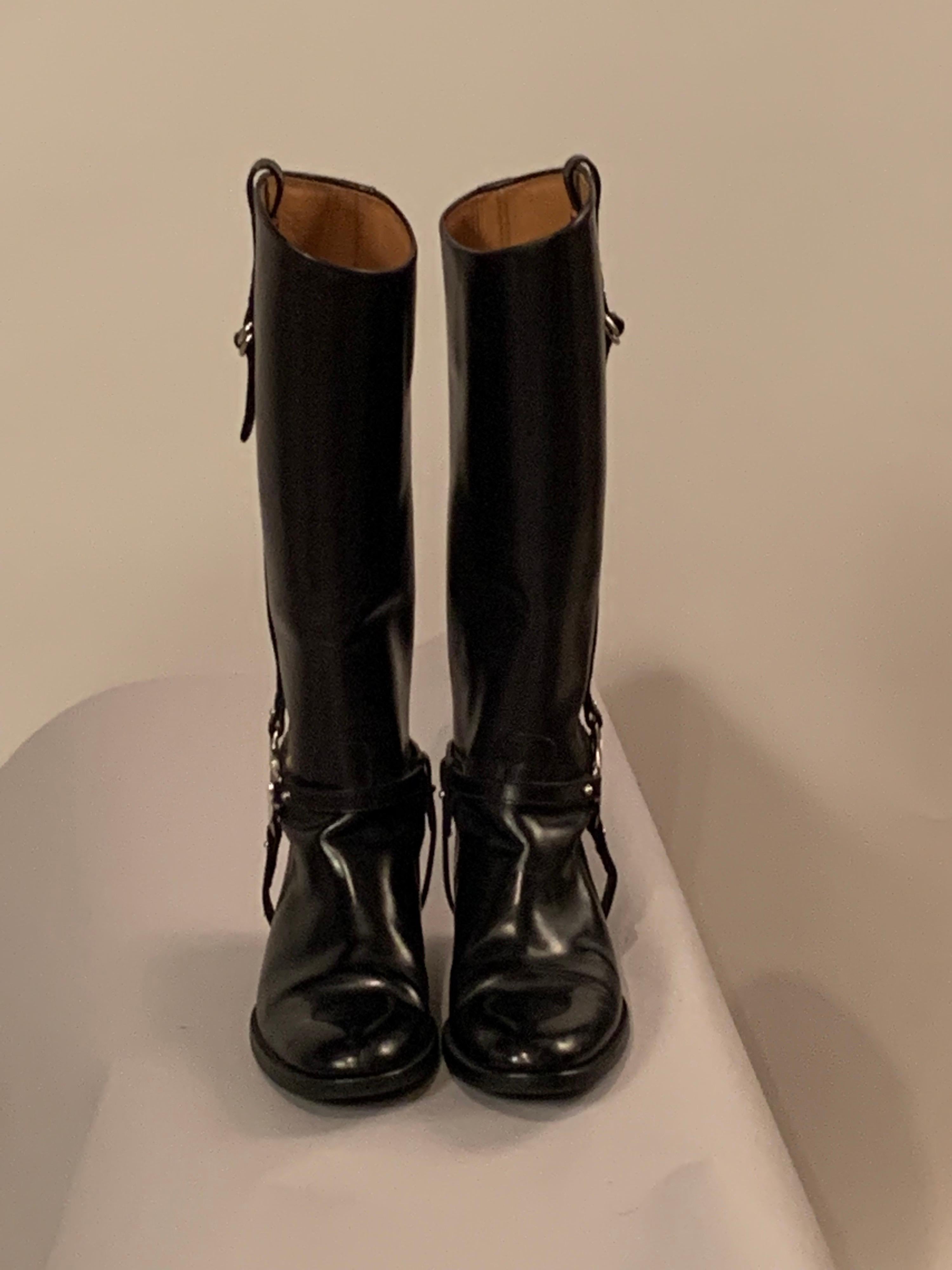 Classic equestrian boot style is used for the design of black leather Gucci boots.  There is a strap and buckle at the top of each boot, and the classic Gucci G logo is used on the straps at the ankle of each boot.  The red and green canvas ribbon