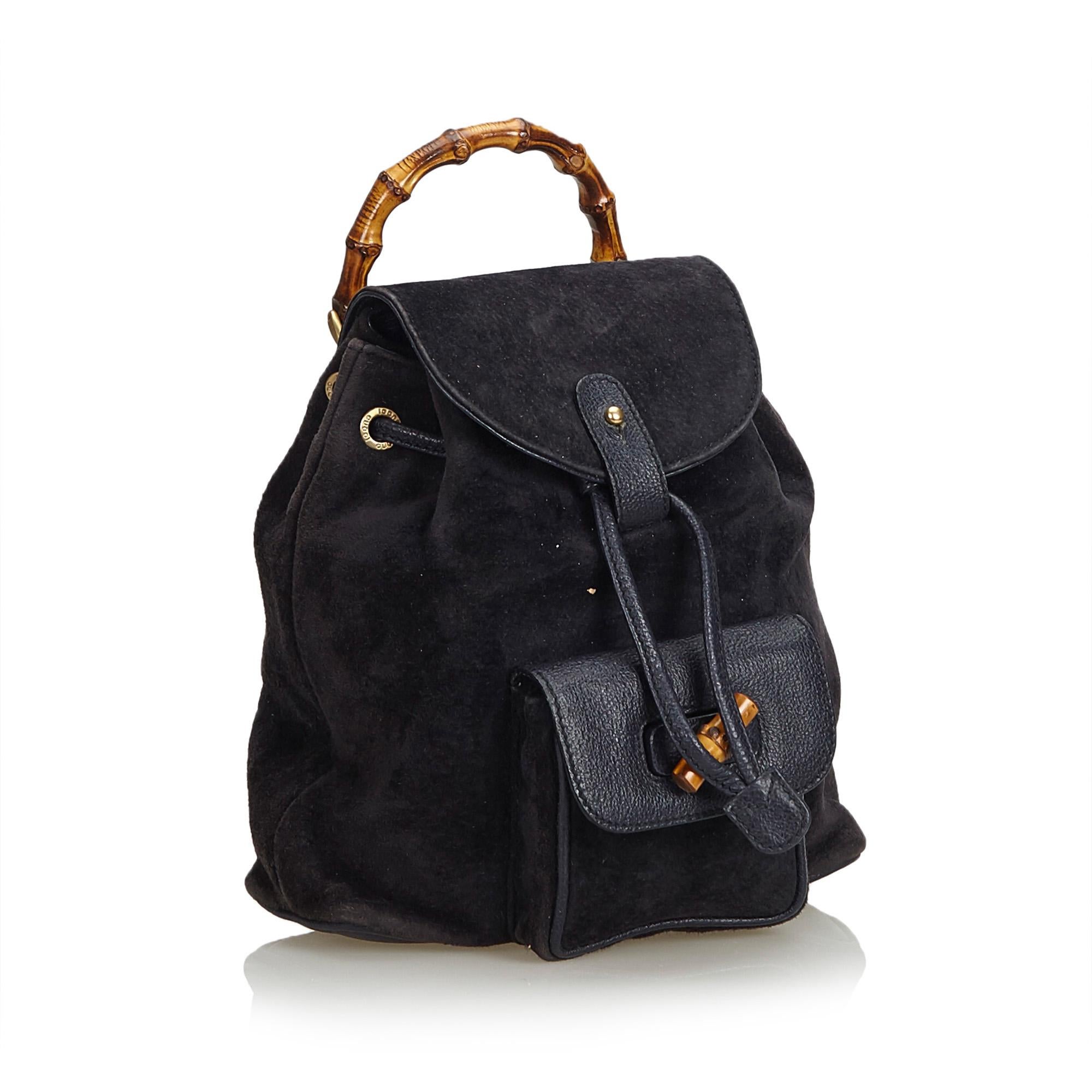 This backpack features a suede body, flat leather back straps, bamboo top handle, top flap with button closure, drawstring closure, exterior flap pocket with bamboo twist lock closure, and interior zip pocket. It carries as B condition