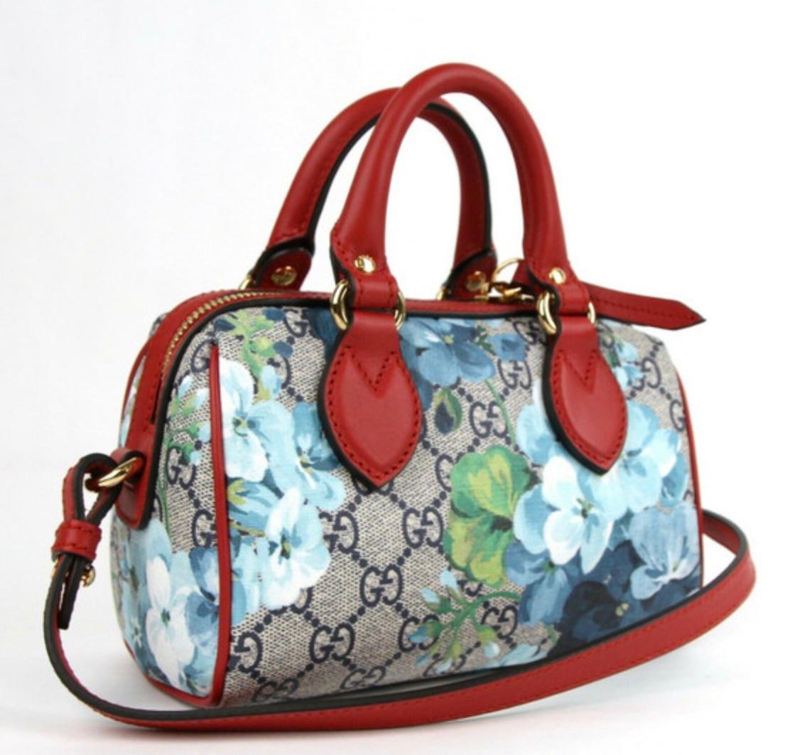 This bag is made with Gucci GG monogram coated canvas in beige with a blue floral print. The handbag features dual rolled leather top handles in red, red trim, an optional red leather shoulder strap, and brass hardware. The top zipper opens to a