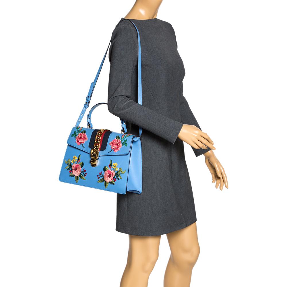 From the house of Gucci comes this gorgeous Sylvie bag that will perfectly complement all your outfits. It has been luxuriously crafted from blue leather and styled with floral embroidery on the front, chain-web decorated flap, and a buckle lock