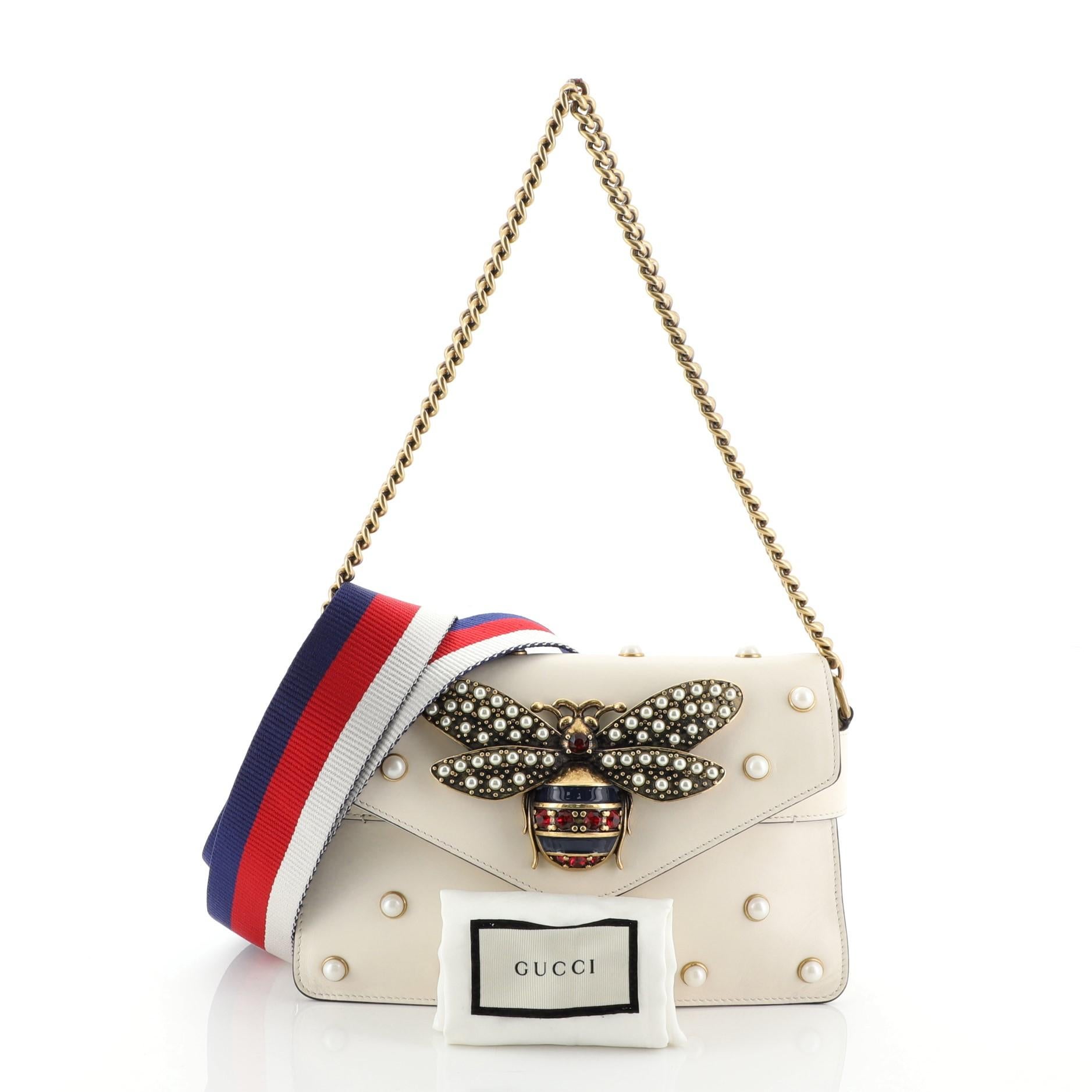 This Gucci Broadway Pearly Bee Shoulder Bag Embellished Leather Mini, crafted from white leather, features chain link strap, multiple pearl studs, metal bee design, and antique brass-tone tone hardware. Its magnetic snap closure opens to a brown