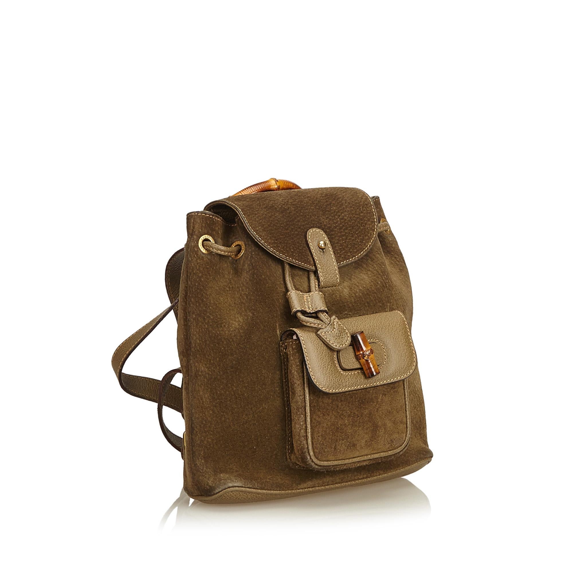 This backpack features a suede body, flat leather back straps, bamboo top handle, top flap with button closure, drawstring closure, exterior flap pocket with bamboo twist lock closure, and an interior zip pocket. It carries as B condition