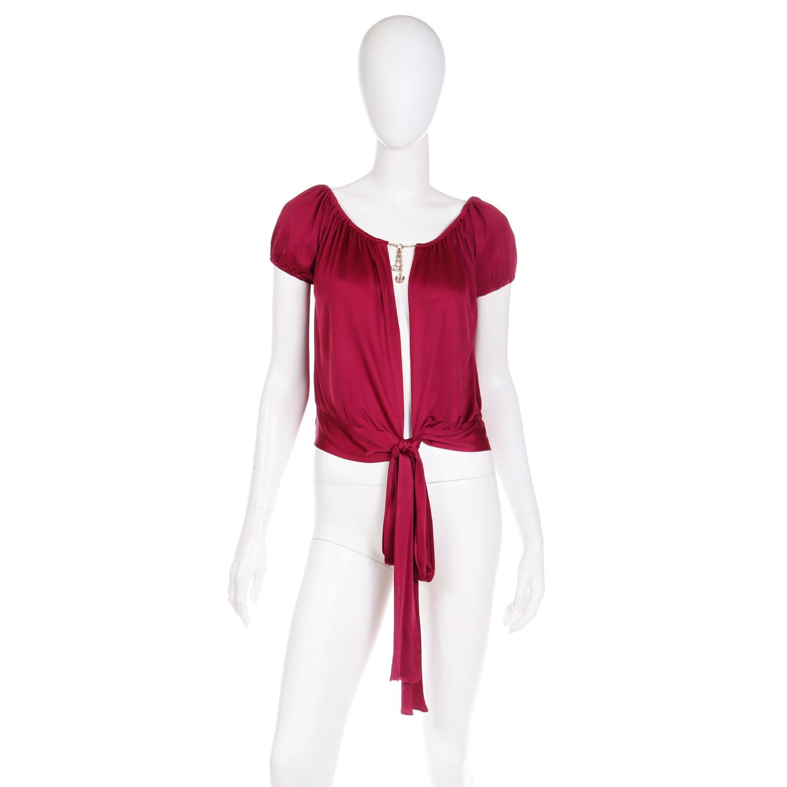 This exquisite burgundy short sleeve Gucci top is a statement piece with its gold GG logo and anchor charms dangling from the chain that connects each side of the neckline. The top is daringly open down the center below the charms and it has a sash