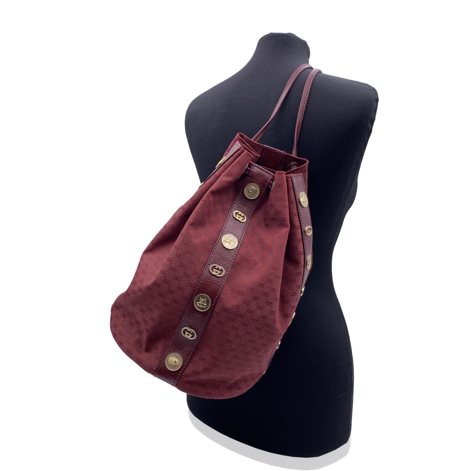 Gucci drawstring bucket backpack in burgundy monogram canvas. The bag is shaped like a bucket bag and embellished with gold metal medallions inspired by a vintage coin that recreates the brand's iconic elements: interlocking G's, feline heads,