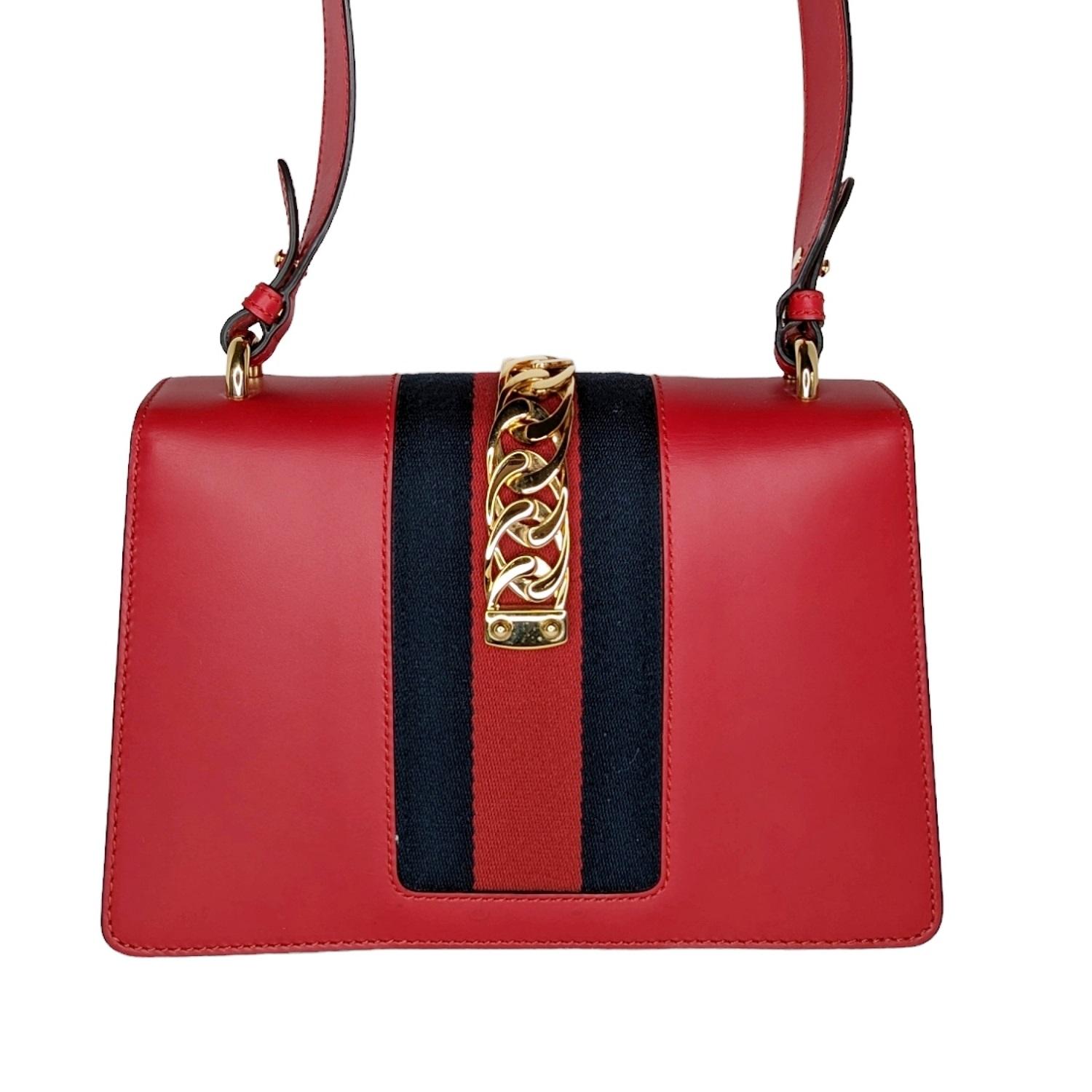 This chic structured shoulder bag is crafted of smooth red calfskin leather and features a prominent front flap detailed with a central canvas red and navy blue web stripe. The bag features a red leather top strap and a shoulder strap of twisted