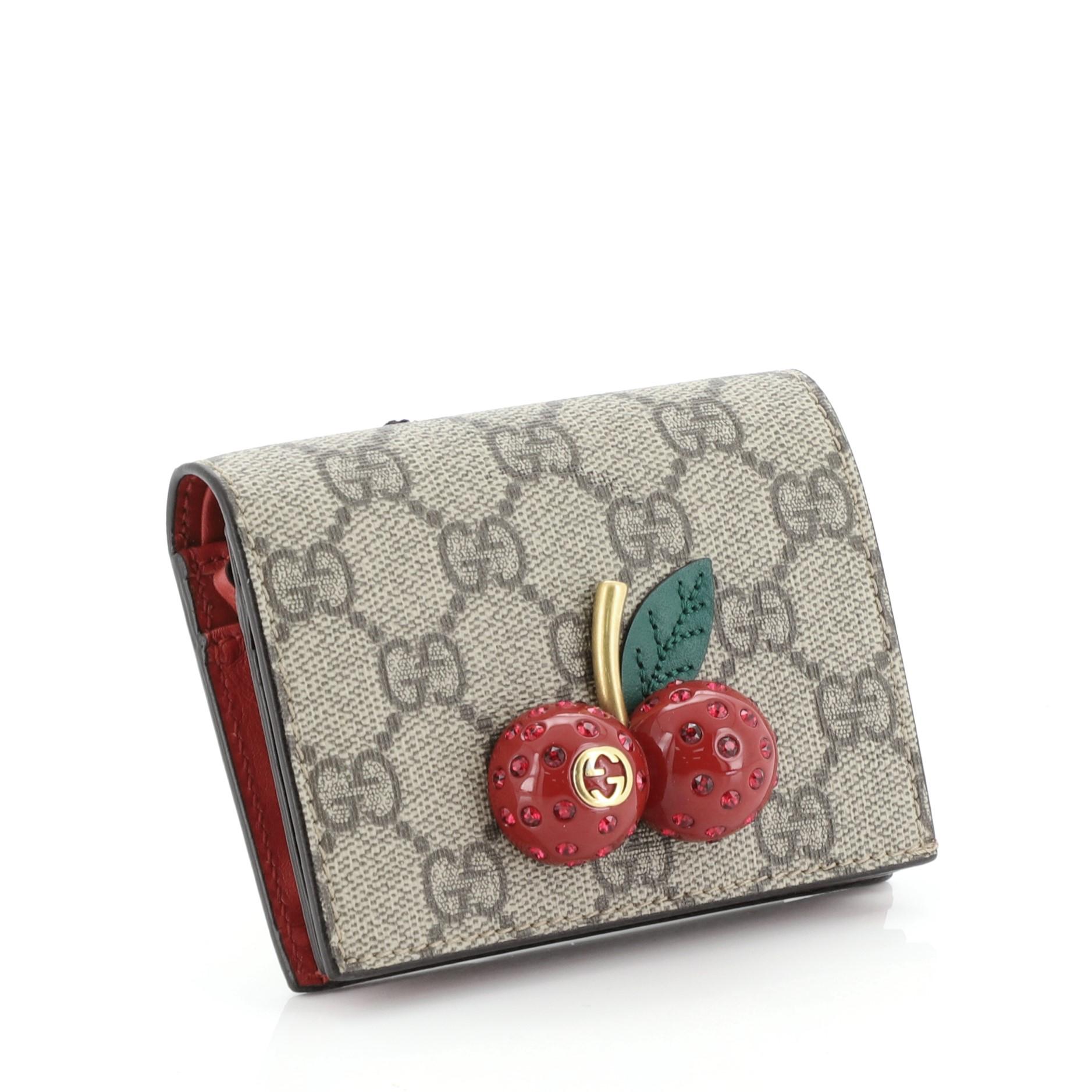 This Gucci Cherries Flap Card Case GG Coated Canvas, crafted from brown GG coated canvas, features 3D jeweled cherry embellishment with a gold stem and interlocking GG logo and aged gold-tone hardware. Its snap button closure opens to a red leather