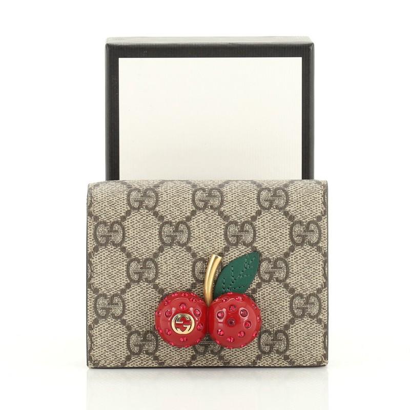This Gucci Cherries Flap Card Case GG Coated Canvas, crafted from brown GG coated canvas, features 3D jeweled cherry embellishment with a gold stem and interlocking GG logo and aged gold-tone hardware. Its snap button closure opens to a red leather
