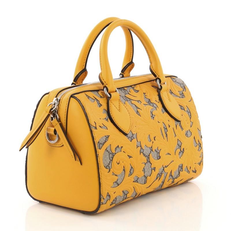 This Gucci Convertible Boston Bag Arabesque GG Coated Canvas Small, crafted from yellow arabesque and brown GG coated canvas, features dual rolled leather handles, overlay of leather floral arabesque pattern, and silver-tone hardware. Its zip