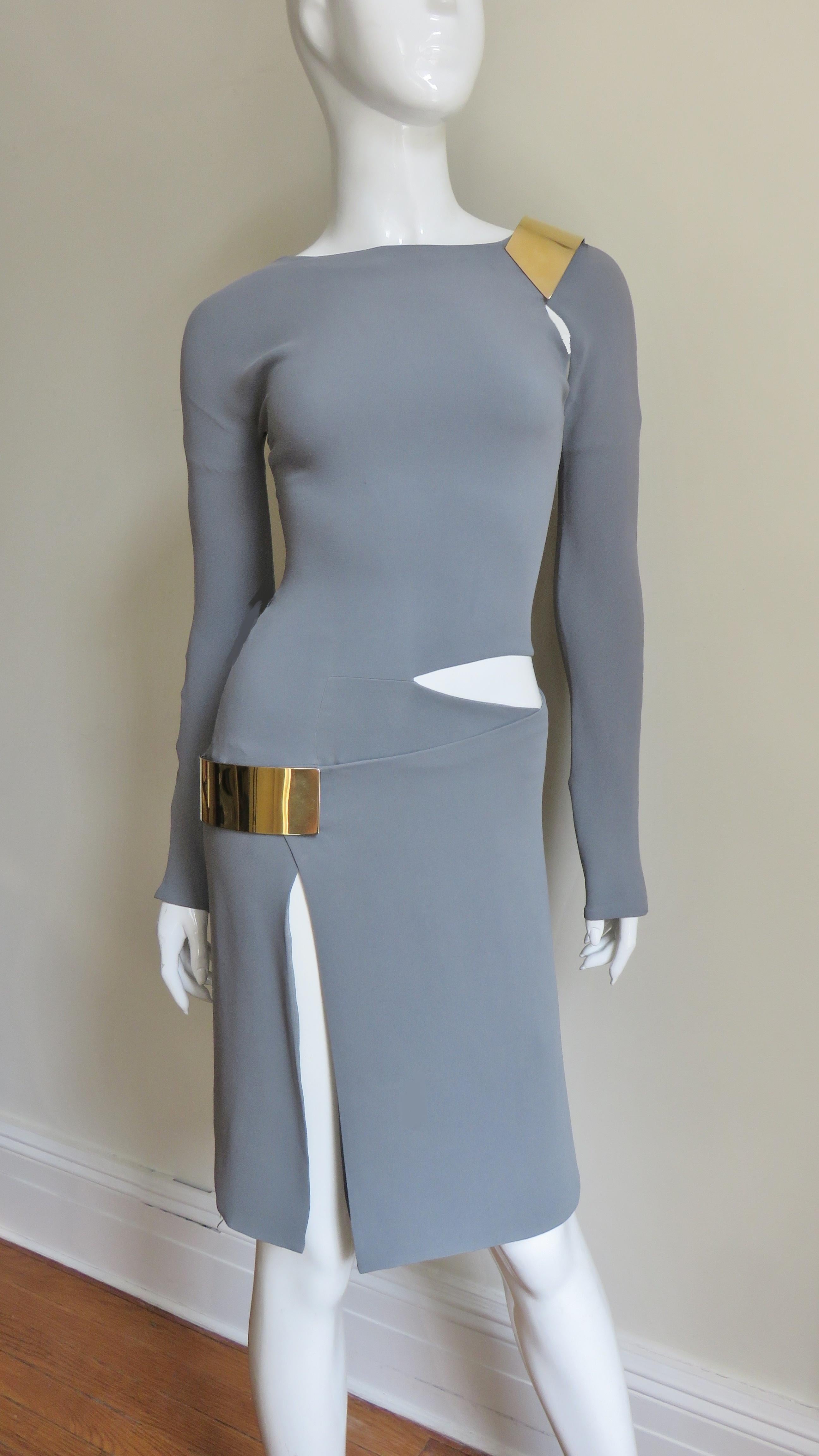 A fabulous grey silk jersey dress from Gucci.  It is fitted with long sleeves and cut outs at one shoulder and side waist plus a slit along the front of one leg of the straight skirt. The slit and shoulder cut out are highlighted with a gold metal