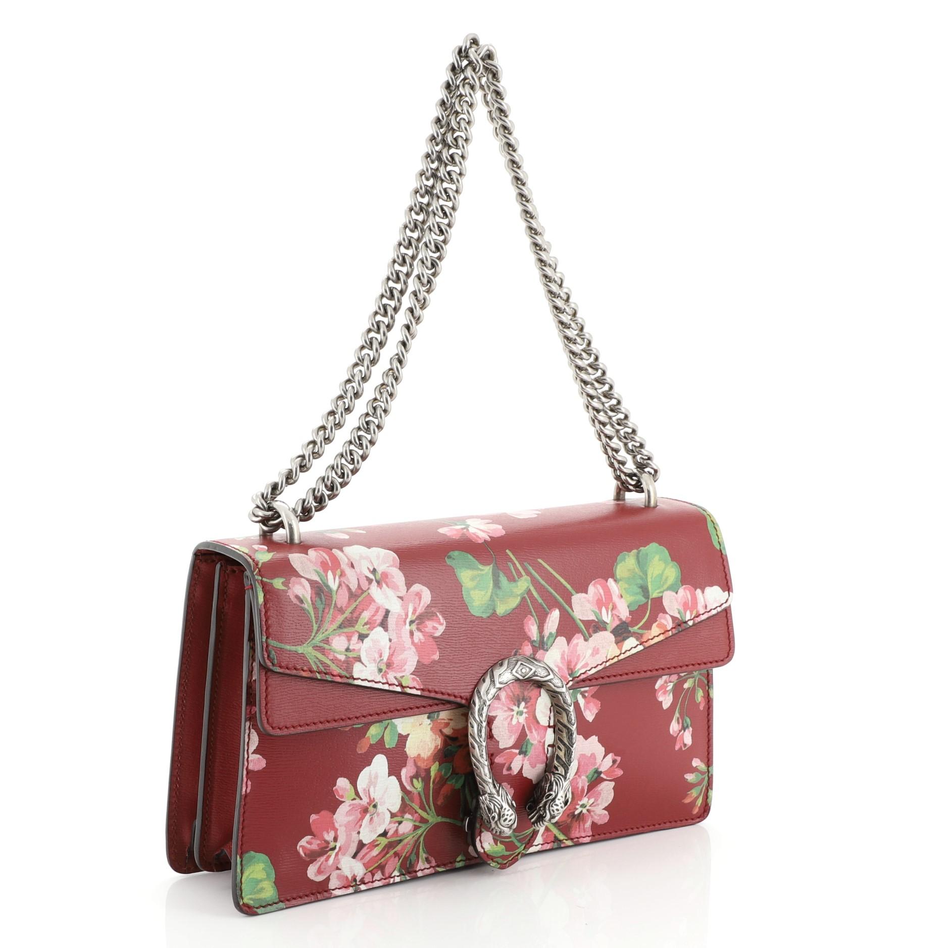 This Gucci Dionysus Bag Blooms Print Leather Small, crafted from red leather with floral print, features sliding chain strap, tiger head spur detail on flap, and aged silver-tone hardware. Its pin closure with side release opens to a neutral canvas