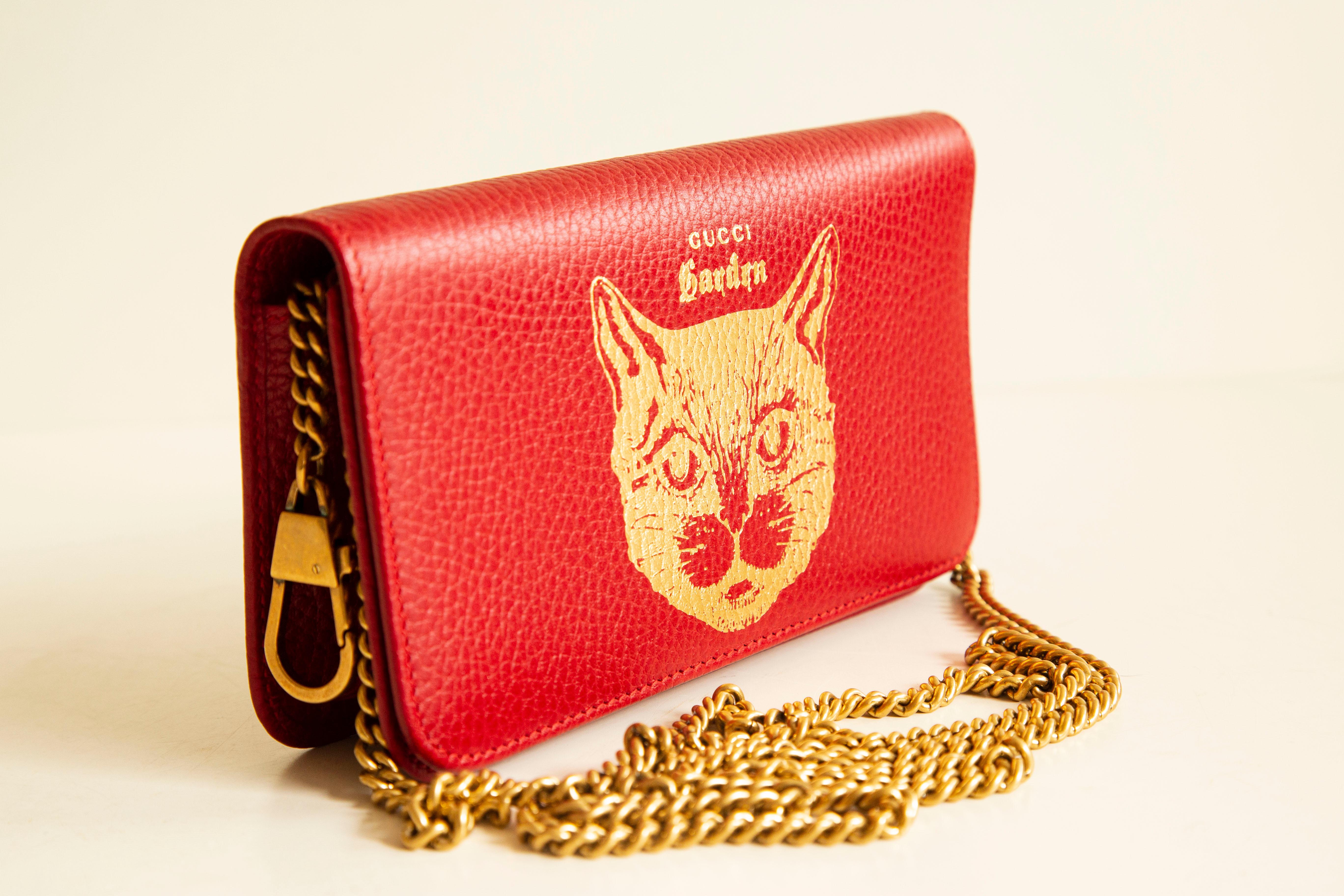 A Gucci limited edition Garden cat chain pouch in red calfskin leather. The bag opens and closes with a magnetic flap and there is one key hanger inside the bag. The interior is lined with blue fabric. The condition is excellent with hardly any