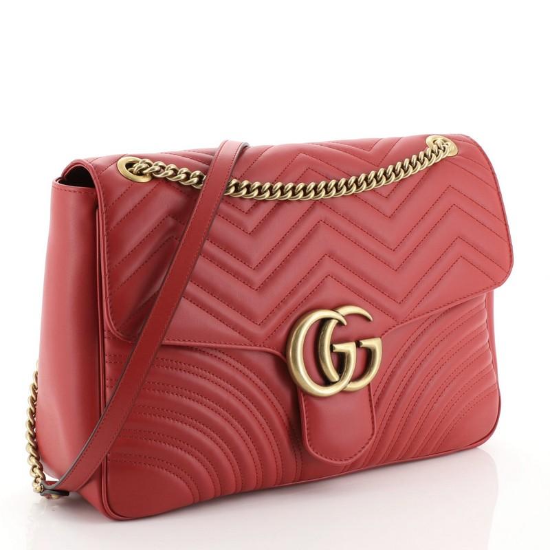 This Gucci GG Marmont Flap Bag Matelasse Leather Large, crafted from red matelasse leather, features a chain-link shoulder strap with leather pad, flap top with the GG logo, and aged gold-tone hardware. Its push-lock closure opens to a neutral