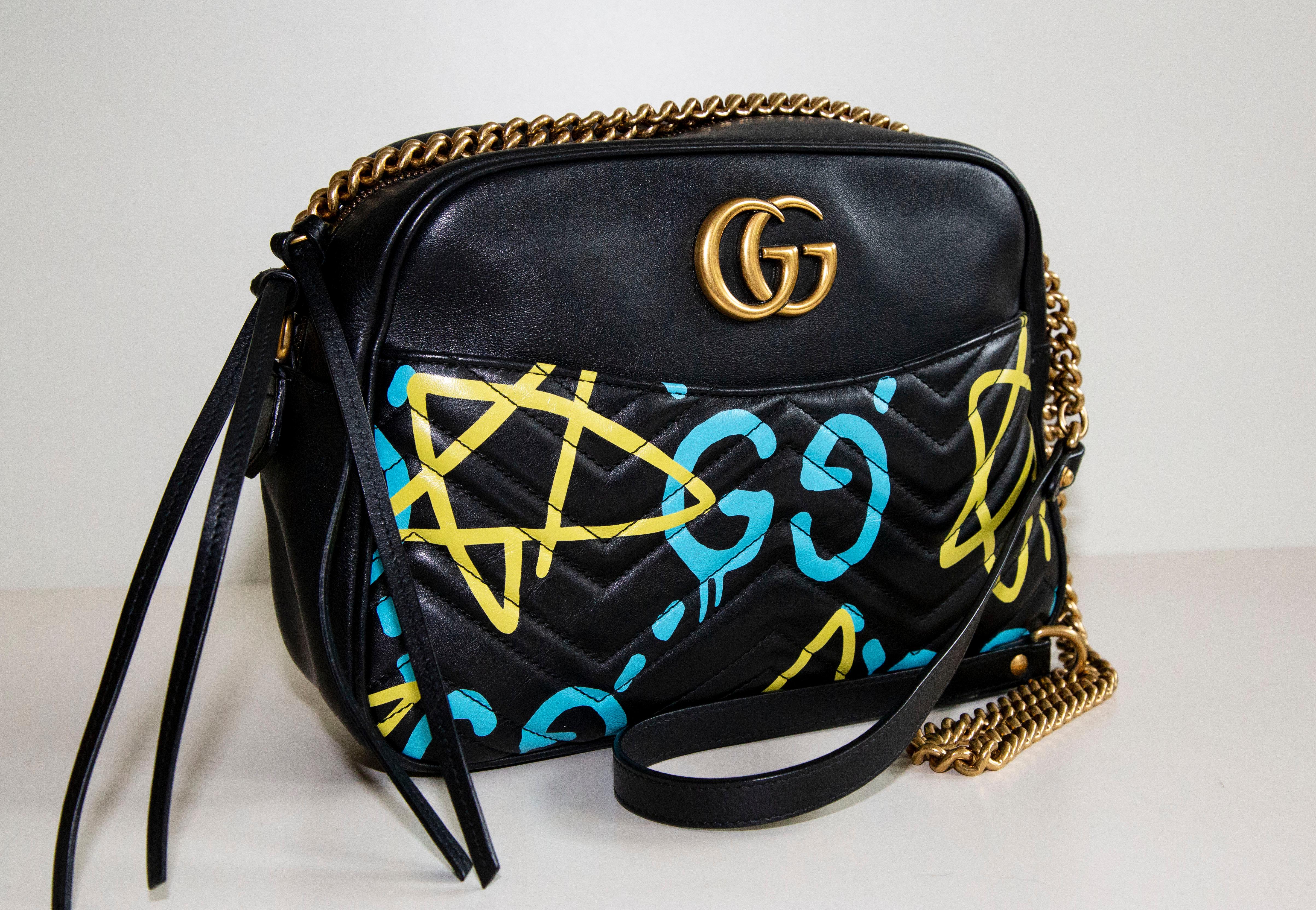 An authentic Gucci Ghost chain crossbody bag. The bag features limited edition spray paint and a handwritten style design. The exterior is made out of soft black leather and gold-tone hardware. The interior is lined with soft beige suede and there