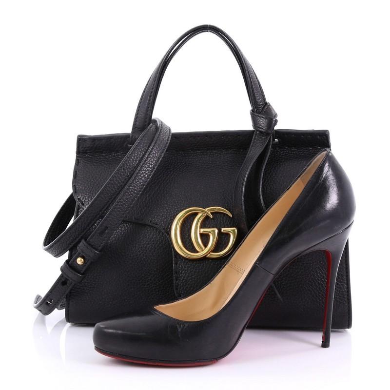This Gucci GG Marmont Top Handle Bag Leather Mini, crafted from black leather, features dual flat handles with knot on one end, flap top with GG logo, top stitching detail, and aged gold-tone hardware. Its push-lock closure underneath the flap opens
