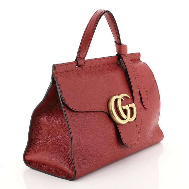 This Gucci GG Marmont Top Handle Bag Leather Small, crafted from red leather, features a flat leather handle, flap top with GG logo, and aged gold-tone hardware. Its push-lock closure opens to a neutral fabric interior with zip and slip pockets.