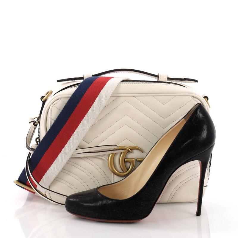 This Gucci GG Marmont Zip Around Camera Bag Matelasse Leather Small, crafted from white matelasse leather, features a flat leather handle, flap top with GG logo, and gold-tone hardware. Its zip around closure opens to a nude microfiber interior with
