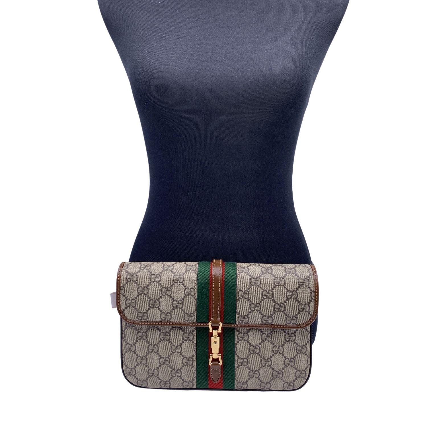 Beige and brown 'Jackie 1961' belt bag from Gucci. Crafted of GG Supreme canvas with brown leather trims. This model is very versatile; it can be used as a belt bag or as a clutch purse if you remove the strap. Logo-engraved snap button, green and