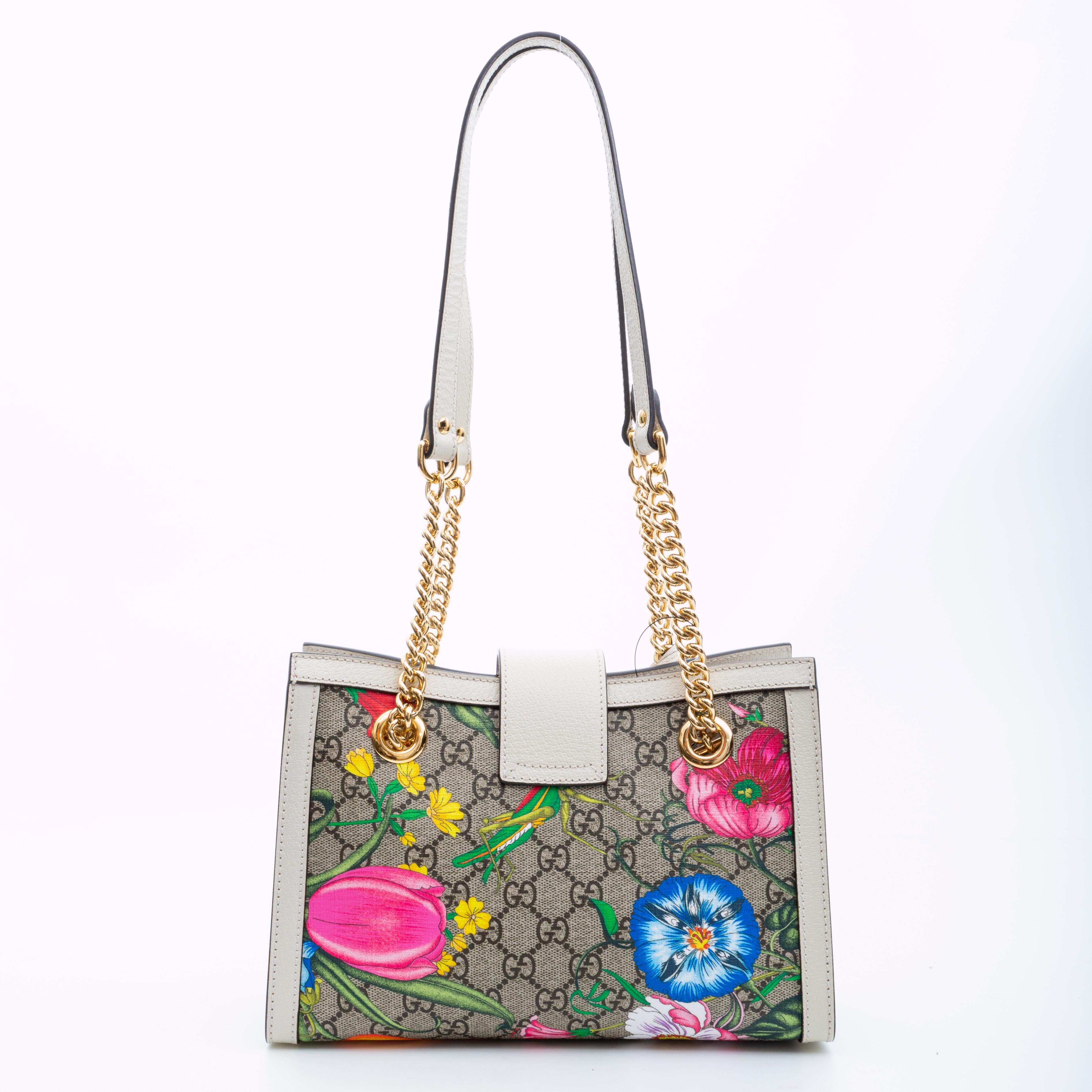 This handbag is made of a GG monogram canvas with a colorful floral printed overlay. The tote features ivory leather trim, polished gold hardware, and dual flat ivory coloured leather and chain shoulder straps. A top crossover leather band with