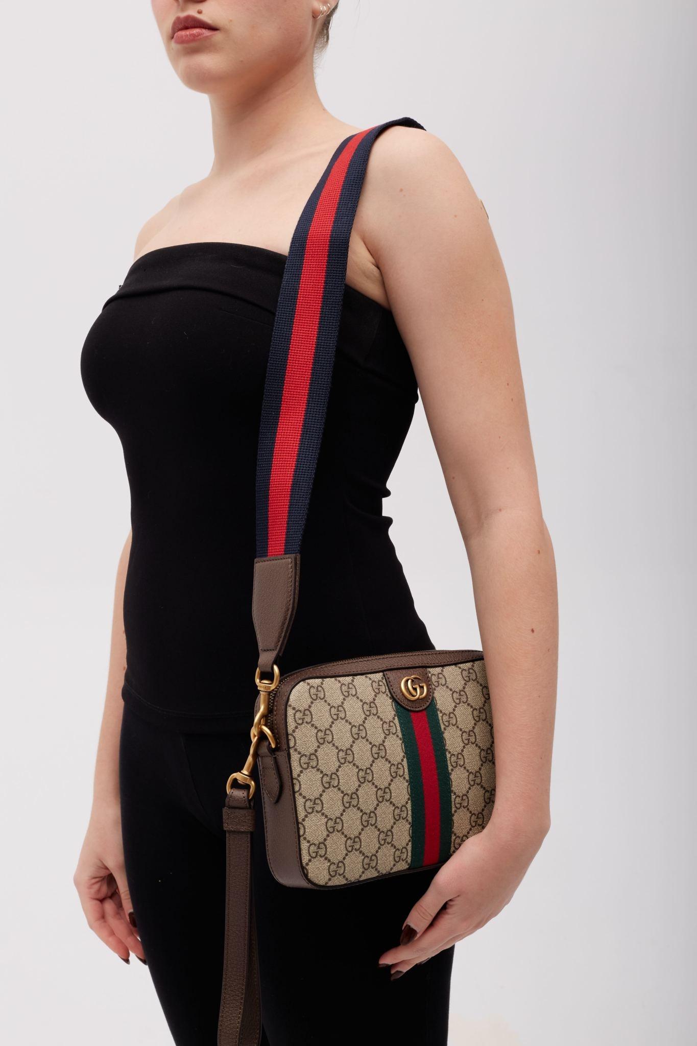 This Gucci bag is made from GG Supreme monogram-coated canvas in an ebony color tone, featuring brown leather accents and a web stripe detail at the front. It is complemented by a red and dark blue web-striped shoulder strap and antiqued gold-tone