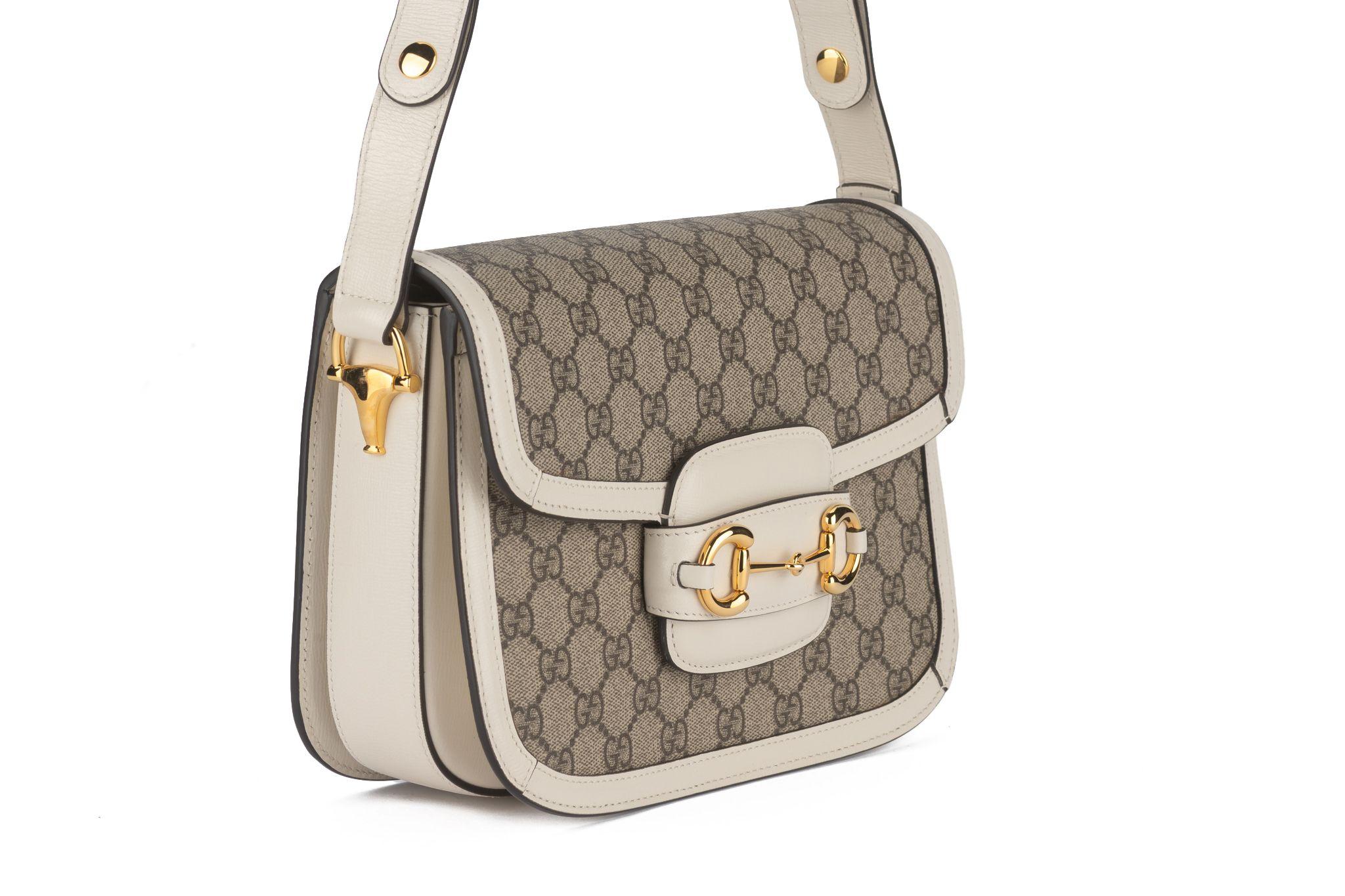 Questa borsa a tracolla Gucci Horsebit 1955 in GG Supreme color crema, con rifiniture in pelle bianca e finiture dorate con un dettaglio a forma di morsetto sul davanti. Ha una tracolla regolabile da 10