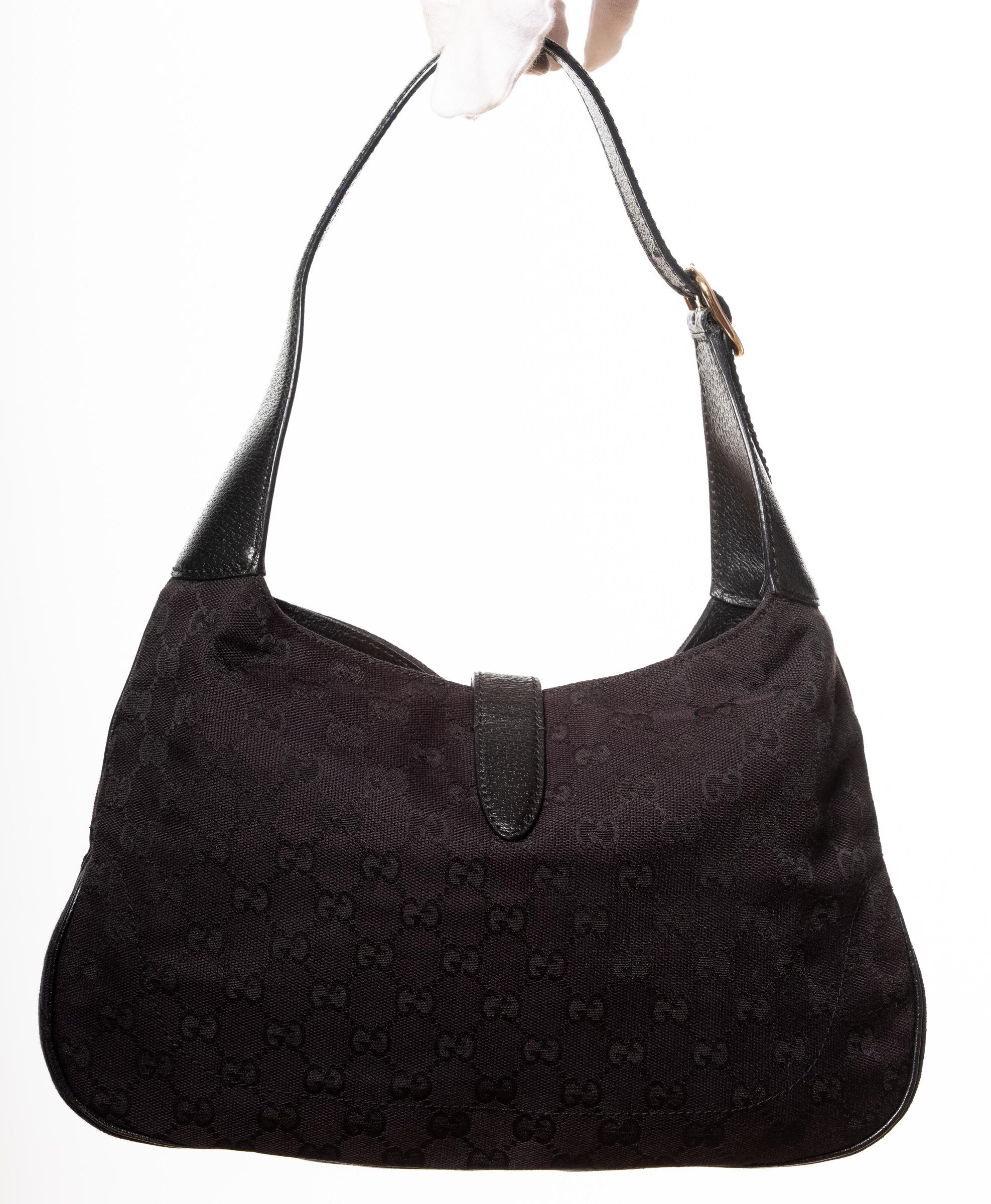 This hobo bag is made of black Gucci GG monogram canvas with leather finishes. The bag features a looping leather shoulder strap and a cross over strap with a rounded silver tone piston lock to secure the top. This opens to a black fabric interior