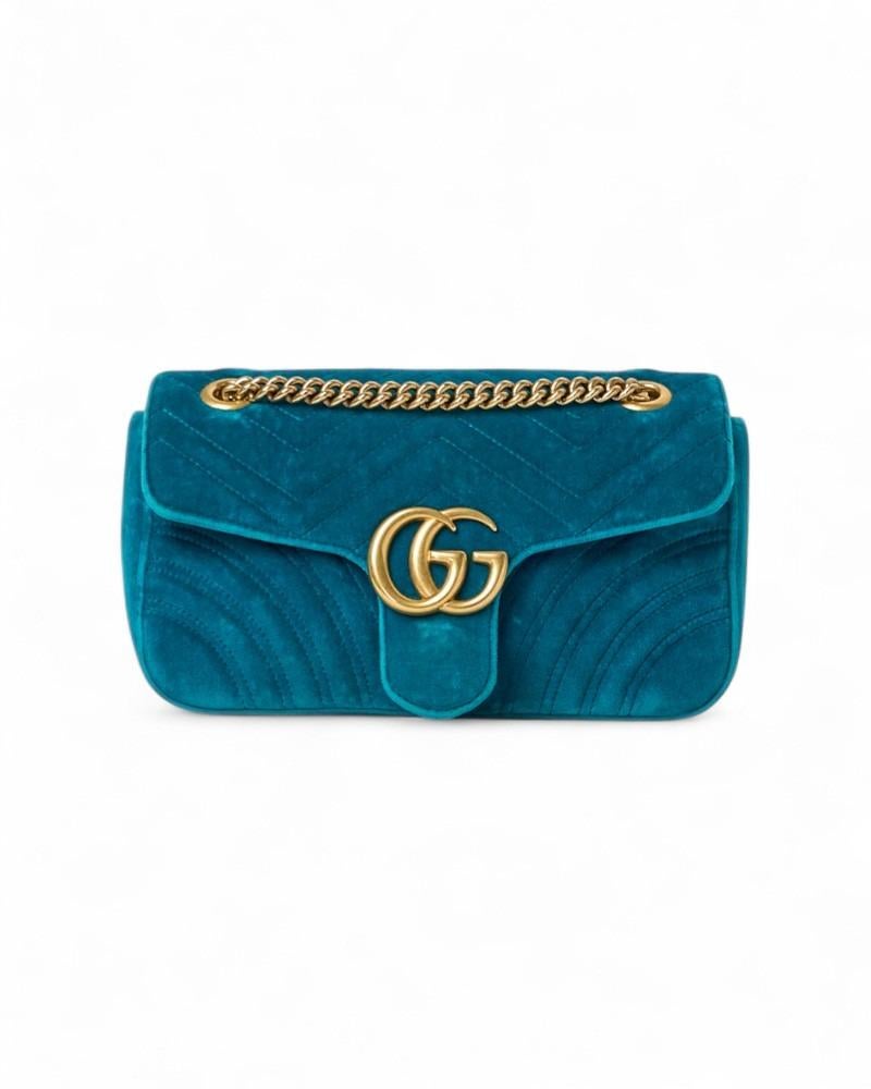 Sac signature de Gucci, ligne Marmont en taille 26, en velours turquoise avec surpiqûres et ferrures dorées. Il est doté d'un rabat sur le devant avec le logo classique de la marque en métal 