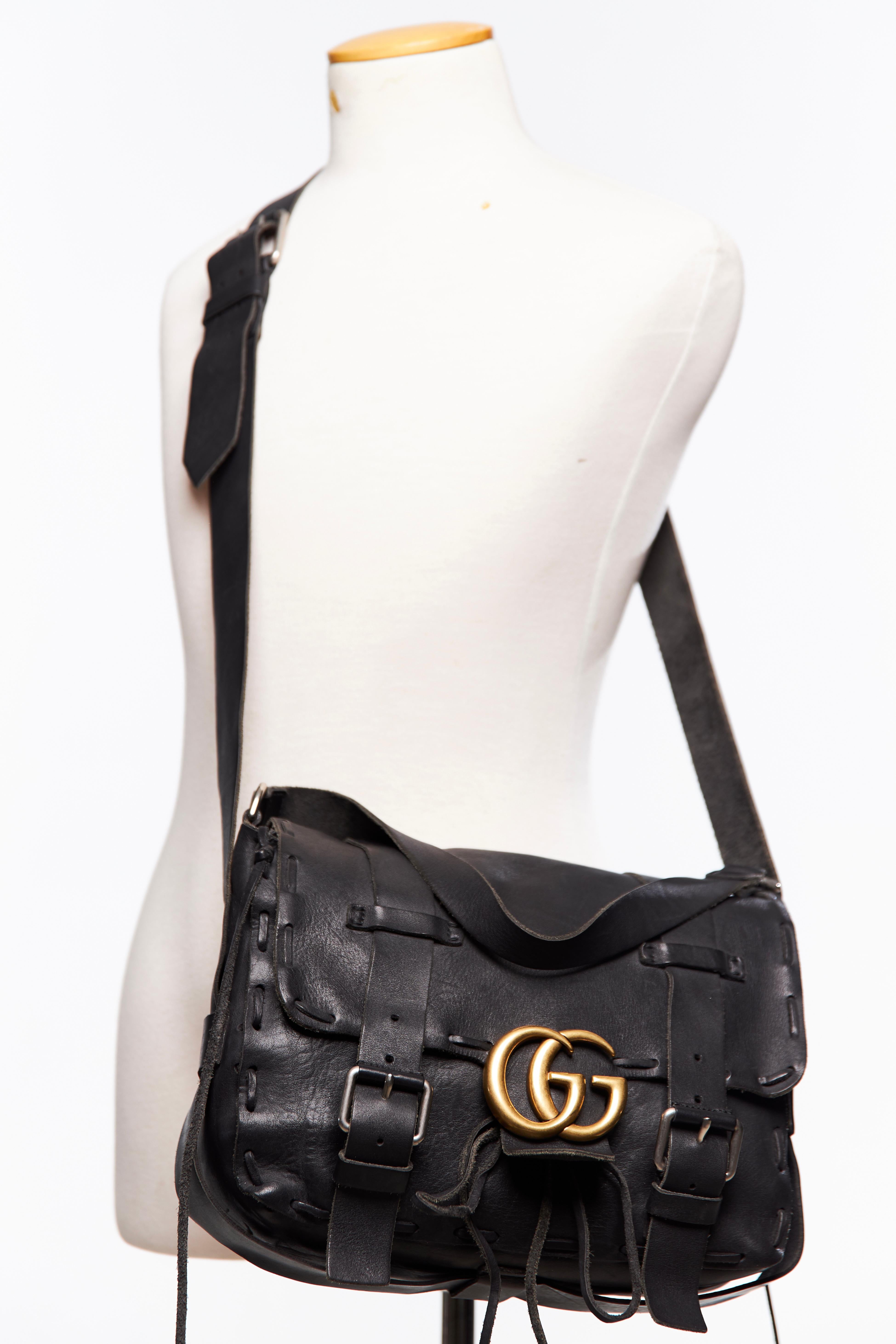 This Gucci messenger bag is made with aged leather in black and features laced edging, an aged gold tone GG, silver hardware, an adjustable shoulder strap, a top handle, black suede interior and a large front flap with buckle and snap