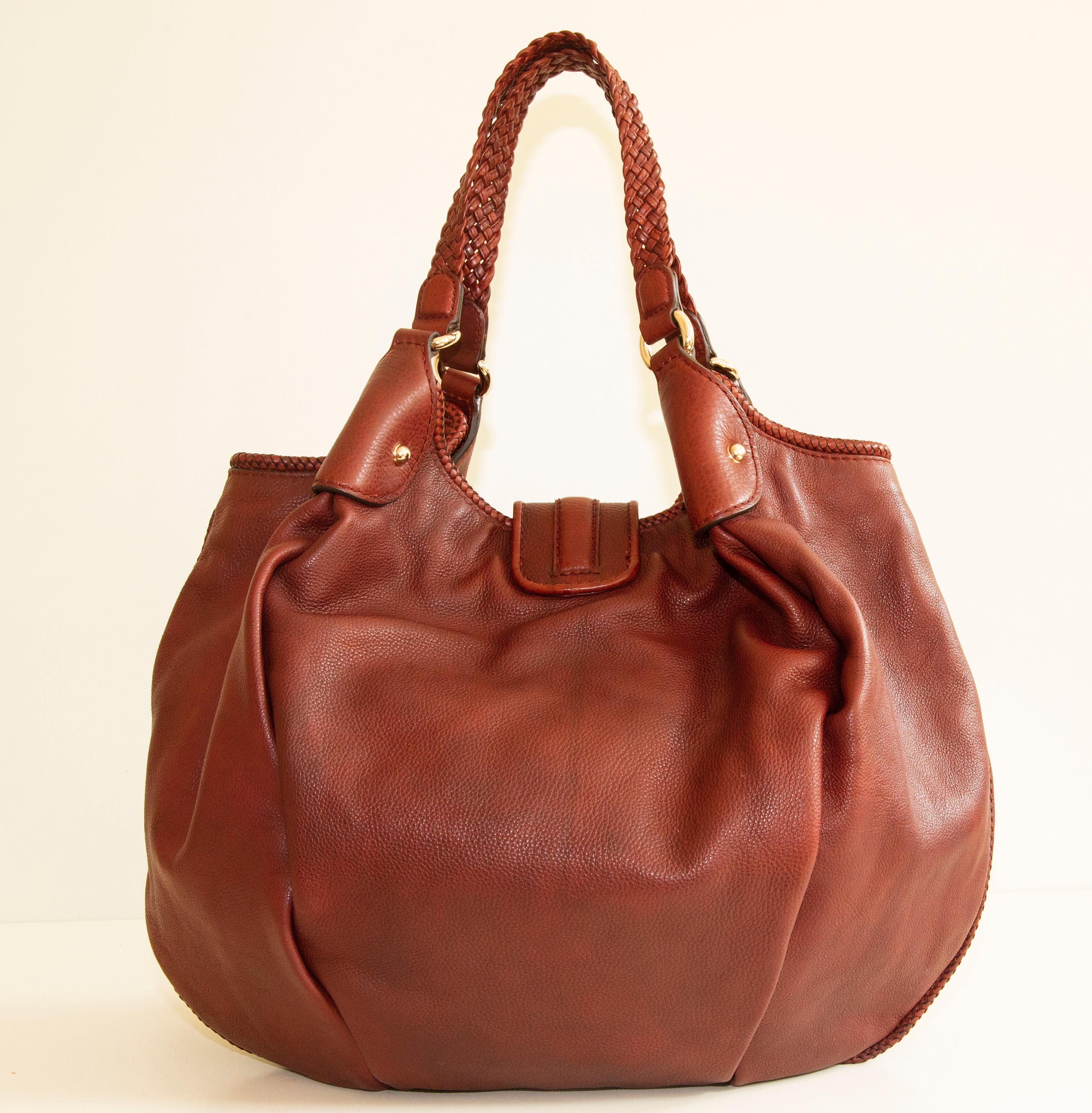 An authentic Gucci Marrakech hobo shoulder bag. The bag features an earth red leather exterior and gold-toned hardware. The interior is lined with beige fabric and there are three side pockets, one zipped pocket, and two slip pockets. The interior