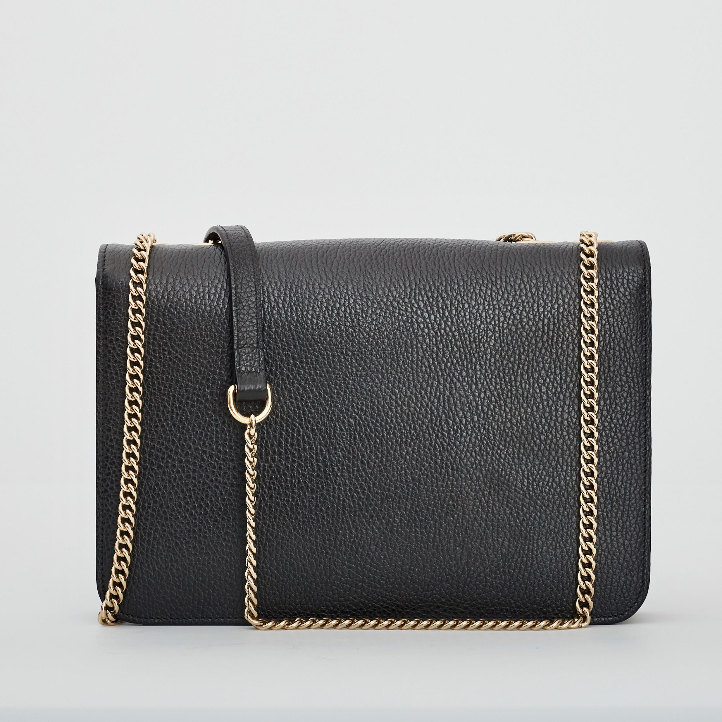 This shoulder bag is made of grained calfskin leather in black. The bag has a waist-length light gold chain shoulder strap and a facing flap with a polished gold interlocking GG emblem lock closure. The flap opens to a partitioned natural canvas