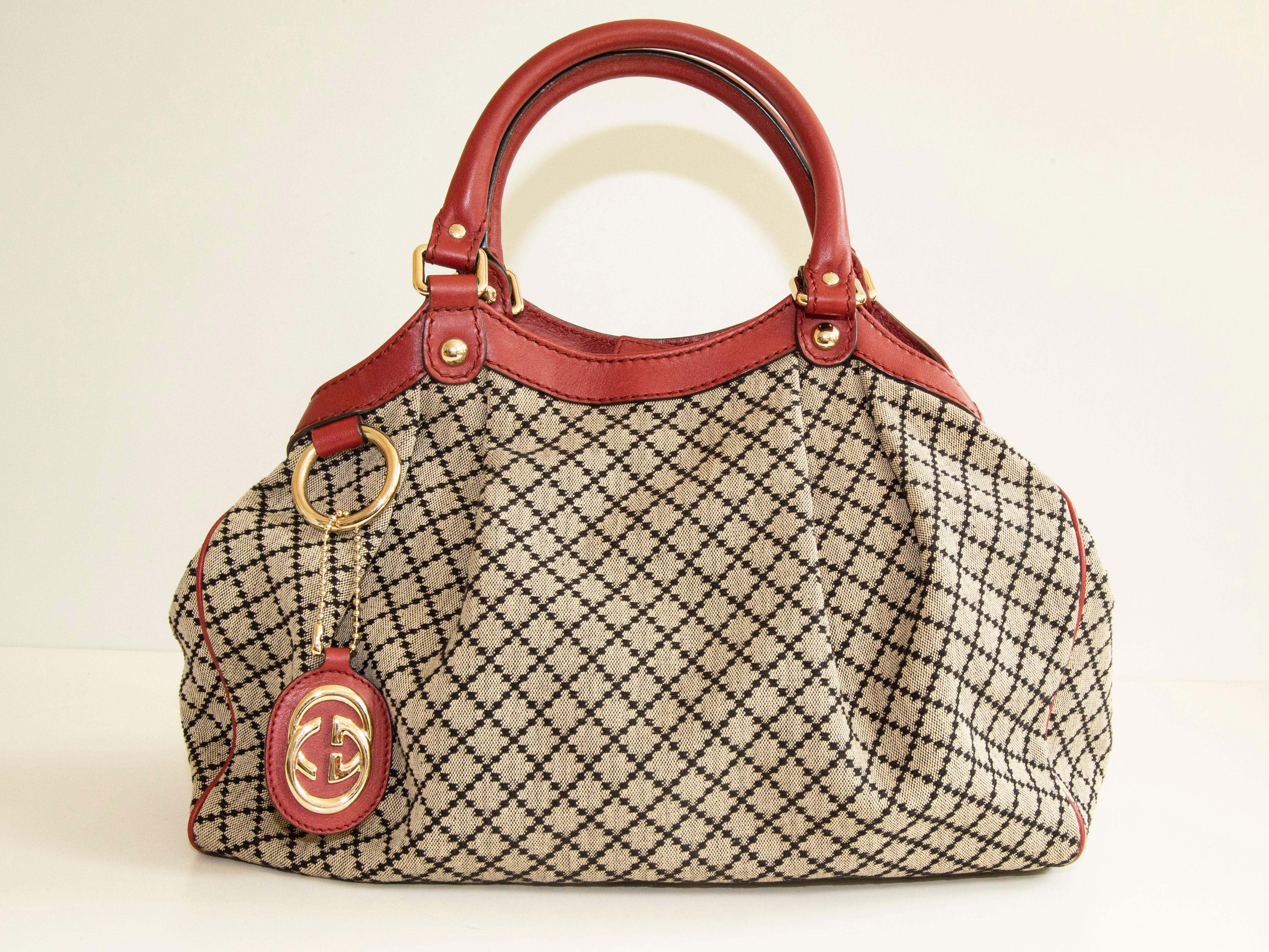 An authentic Gucci Sukey medium handbag/tote bag. The bag features a Diamante canvas, red leather trim, and light-gold-toned hardware. The interior is lined with brown fabric and there is one zipped side pocket. The interior is neat & clean. The