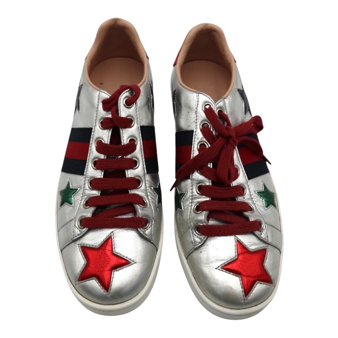 Gucci Metallic Silver Ace Sneakers with Stars & Web Stripe

A bold twist on a classic, these Gucci Ace low-top sneakers feature a metallic silver leather base adorned with signature red and green stars, red laces, and the iconic web stripe in navy
