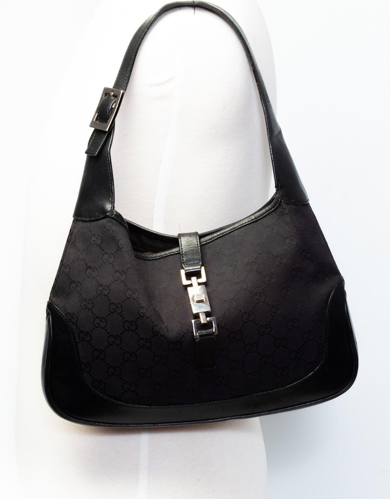 This Gucci Jackie bag is made of black gucci GG monogram canvas with leather finishes. The bag features a looping leather shoulder strap, gun metal hardware, a cross over strap and piston lock closure. The top opens to a black fabric interior with