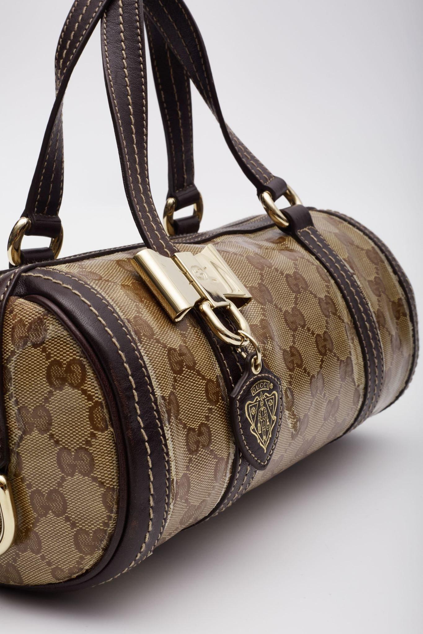 Ce sac Boston est confectionné en toile enduite brillante au monogramme GG de Gucci, avec un fini glacé. Le sac présente des anses en bandoulière, une garniture en cuir marron chocolat avec des maillons en or poli, un nœud décoratif en laiton poli