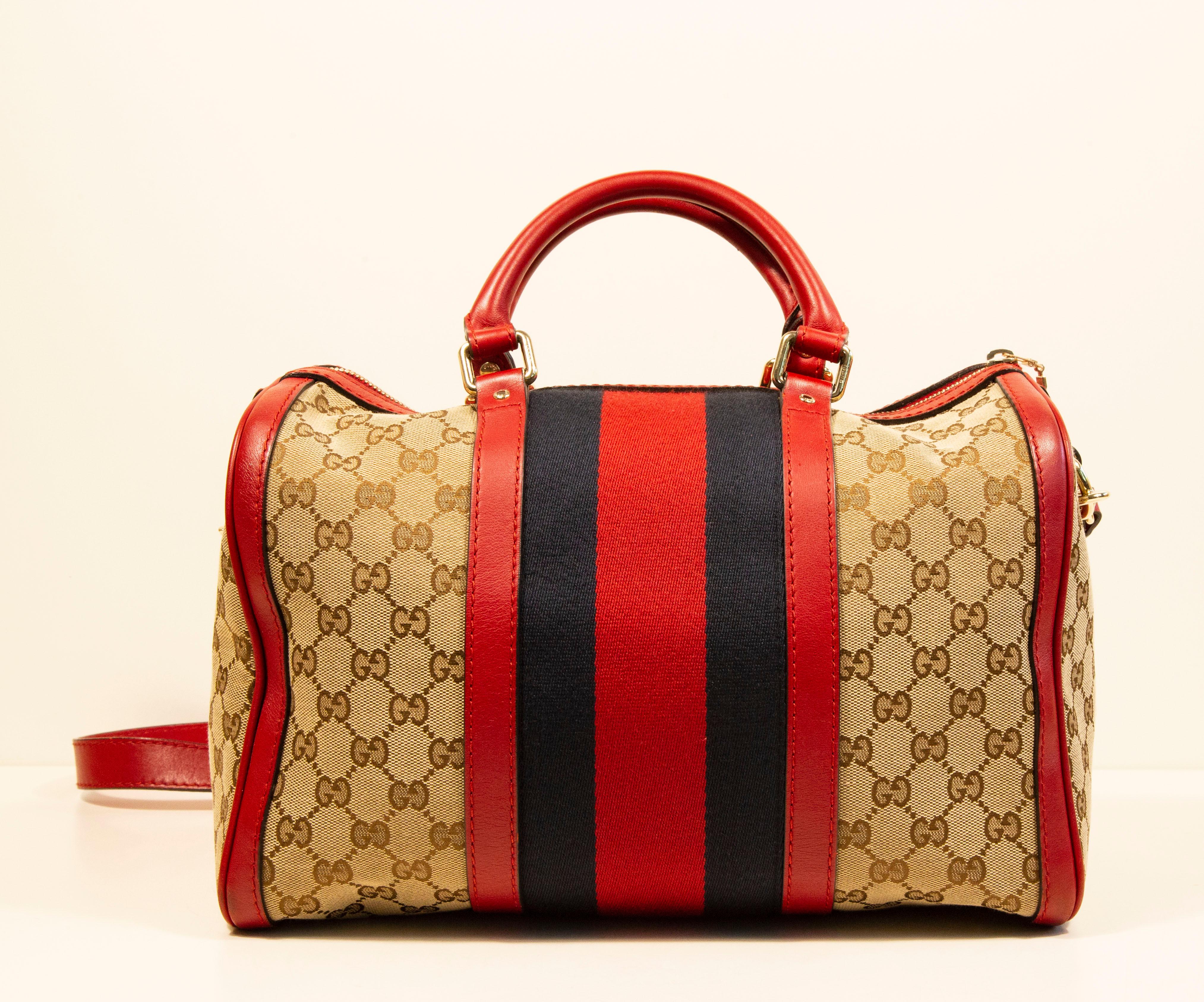 Gucci Boston bag with a shoulder strap. The bag is made of GG brown/beige canvas and red leather trim. The bag features a navy/red Gucci signature race strap. The hardware is gold toned.  The  interior is lined with beige fabric, and next to the