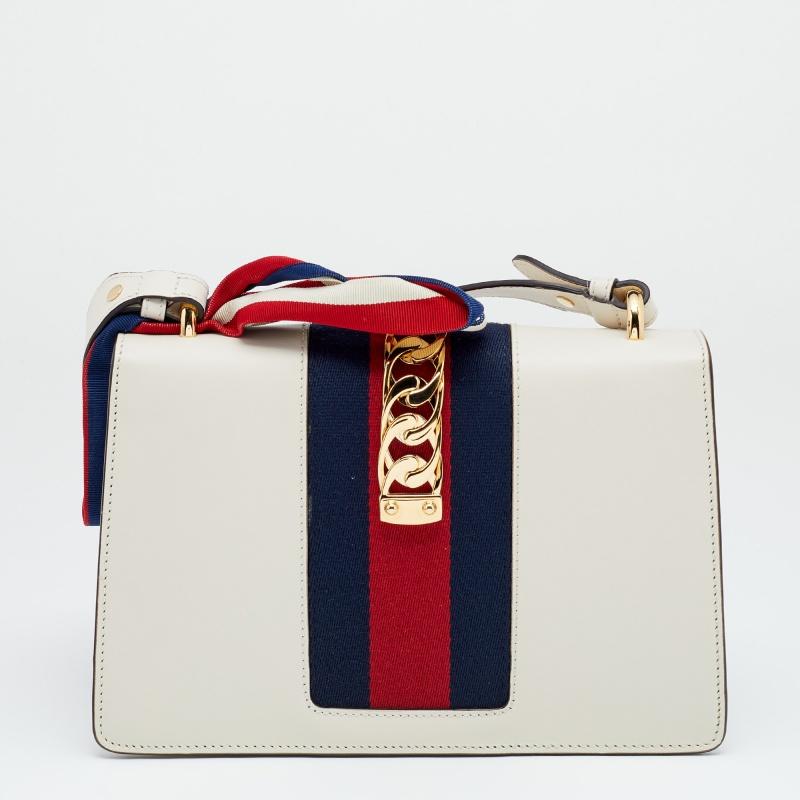 All the designs from Gucci, like this Sylvie bag, reflect a sense of innovation and tradition. Crafted from leather, it is admired for its timeless elegance and structured silhouette. The off-white exterior of this bag is decorated with eye-catching