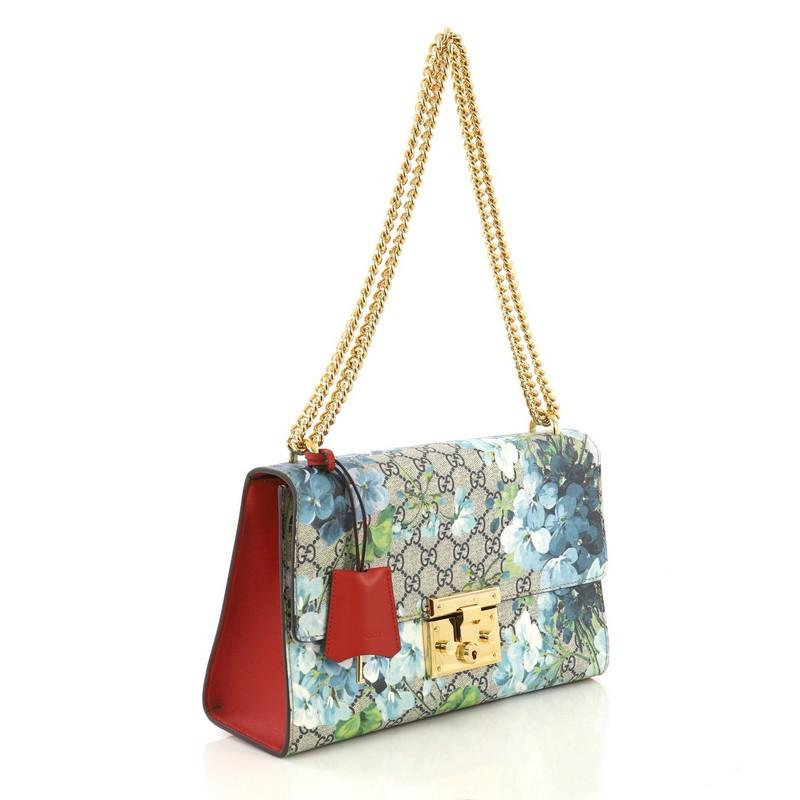 This Gucci Padlock Shoulder Bag Blooms Print GG Coated Canvas Medium, crafted in blue blooms print GG coated canvas and red leather, features chain link strap, exterior back pocket, and gold-tone hardware. Its push-lock closure opens to a light pink