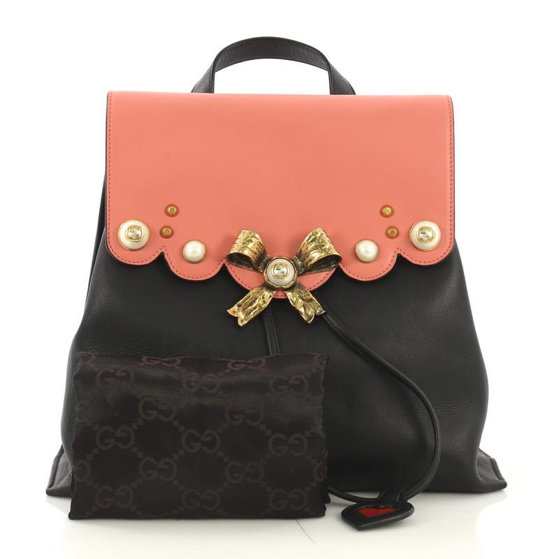 This Gucci Pearly Peony Backpack Leather Medium, crafted from pink and black leather, features adjustable chain shoulder straps, bow accent with pearls, and aged gold-tone hardware. Its flap with magnetic closure opens to a beige fabric interior